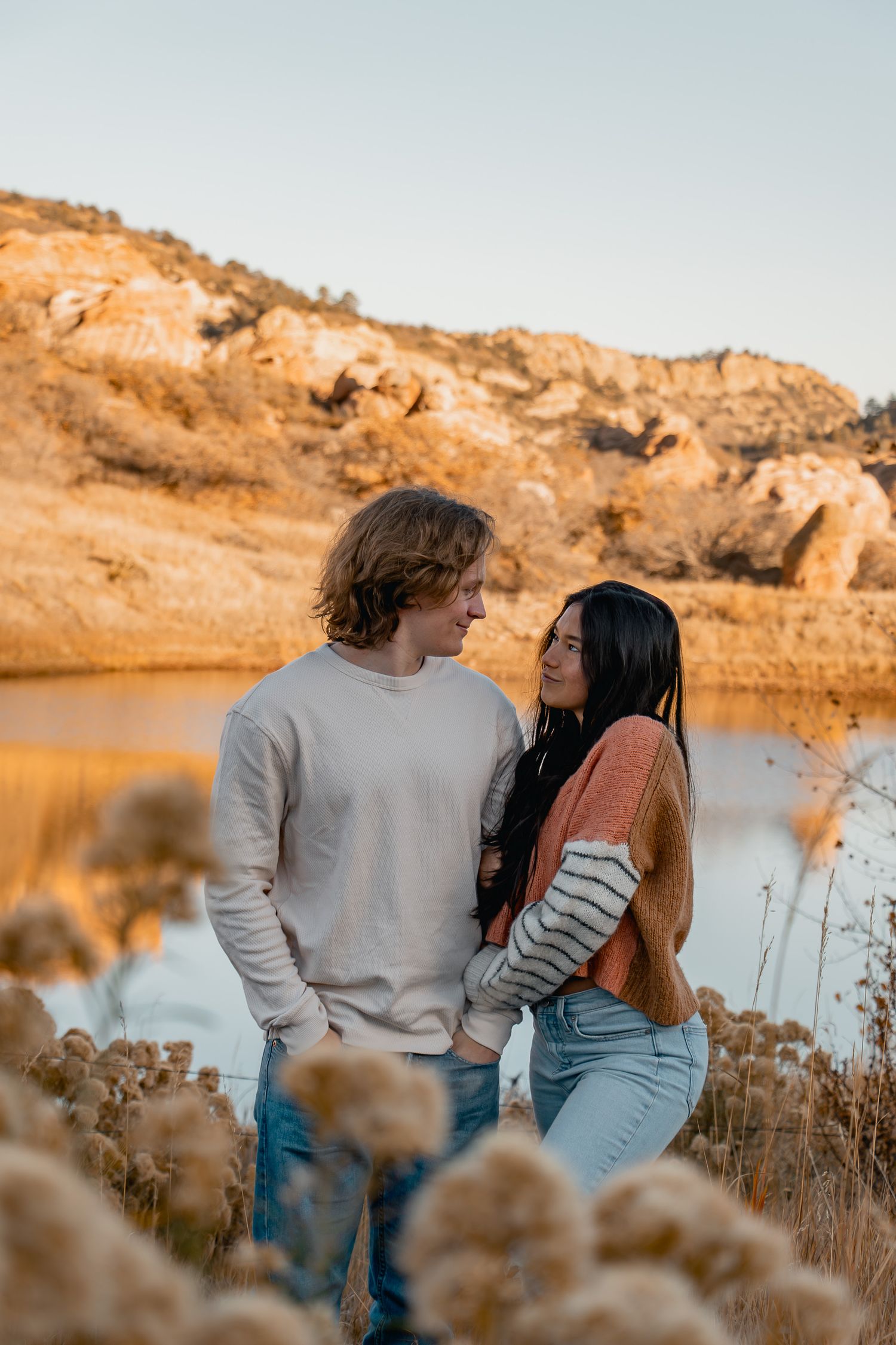 A couple stands by a serene lake at sunset with golden cliffs in the background, creating a romantic silhouette.