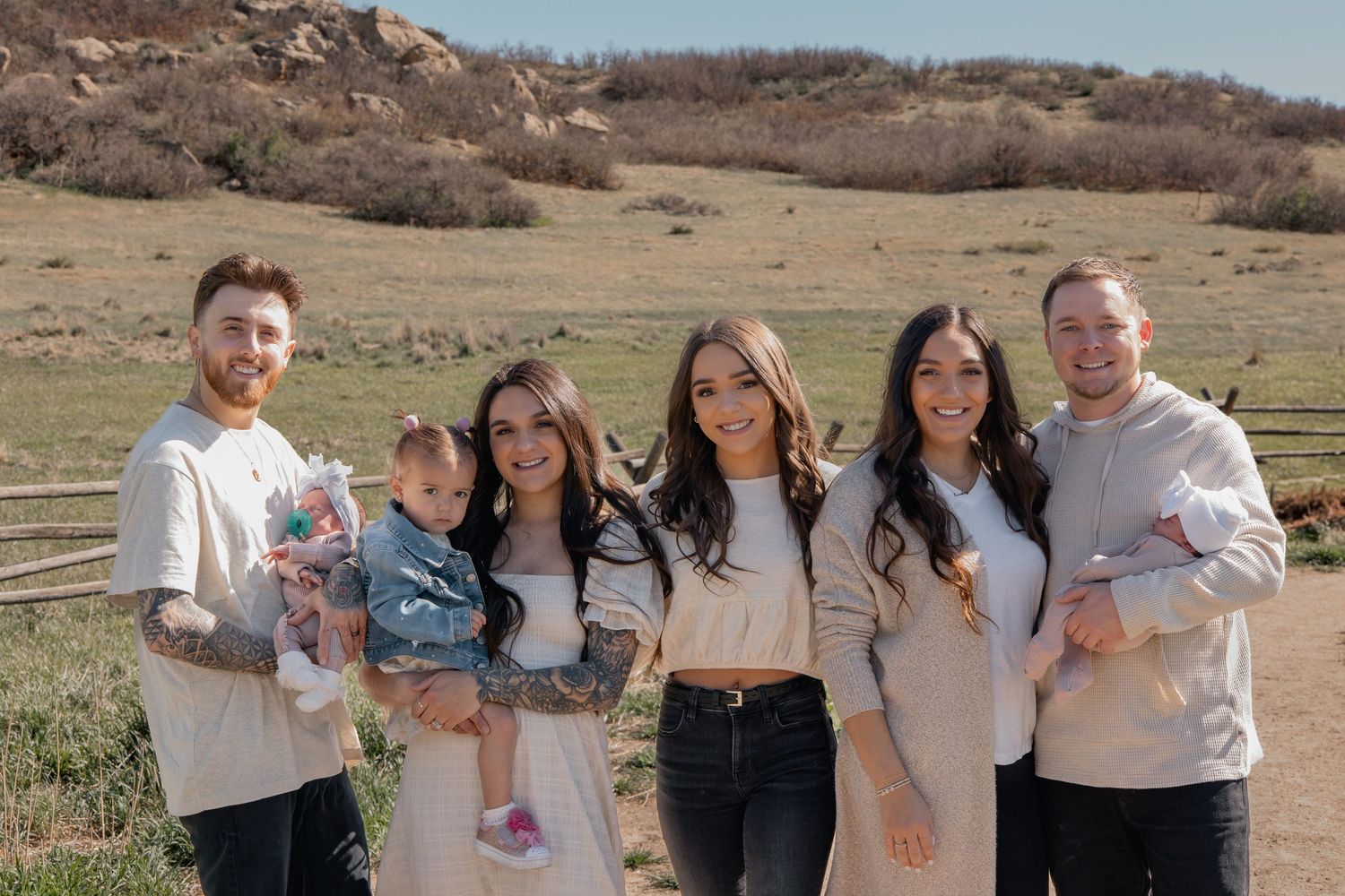 A group stands together outdoors on a sunny day in neutral-colored clothing against a natural hillside backdrop.