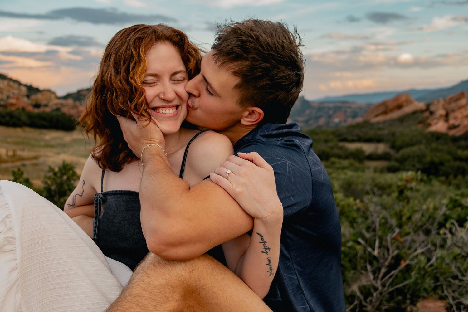 A couple shares a tender moment while embracing in front of a scenic mountainous landscape at sunset.