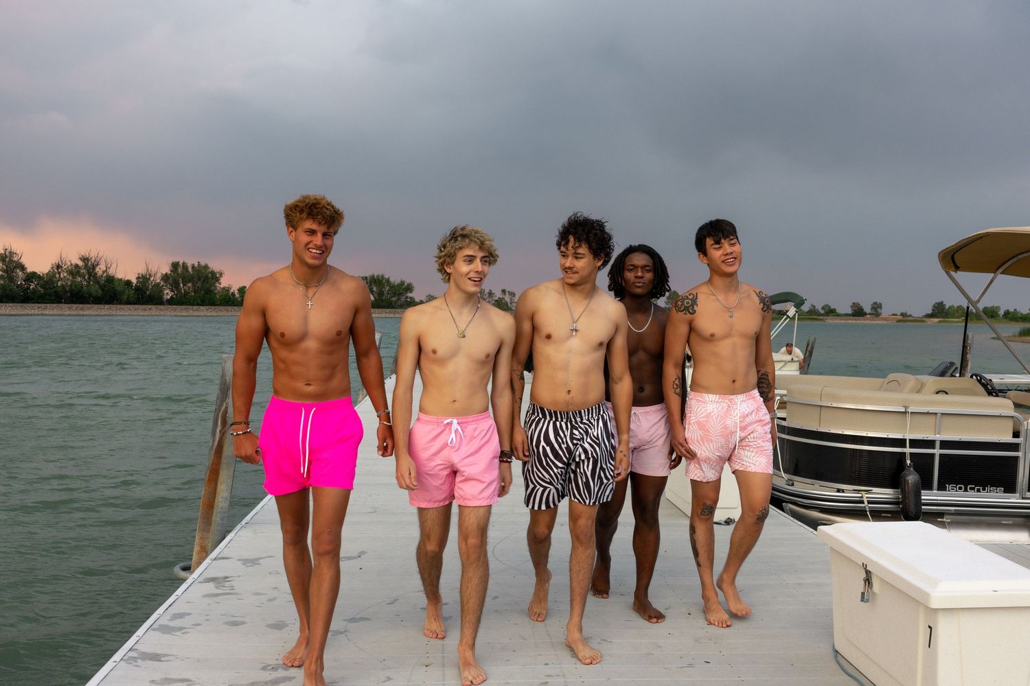 Five shirtless teen boys in colorful swim trunks walk on a dock by a lake under a dramatic cloudy sky.