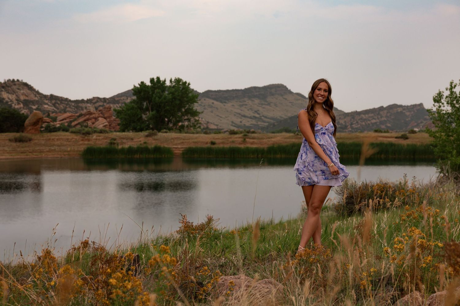 Young woman in floral dress posing by a serene lake with rocky hills and lush greenery at dusk.