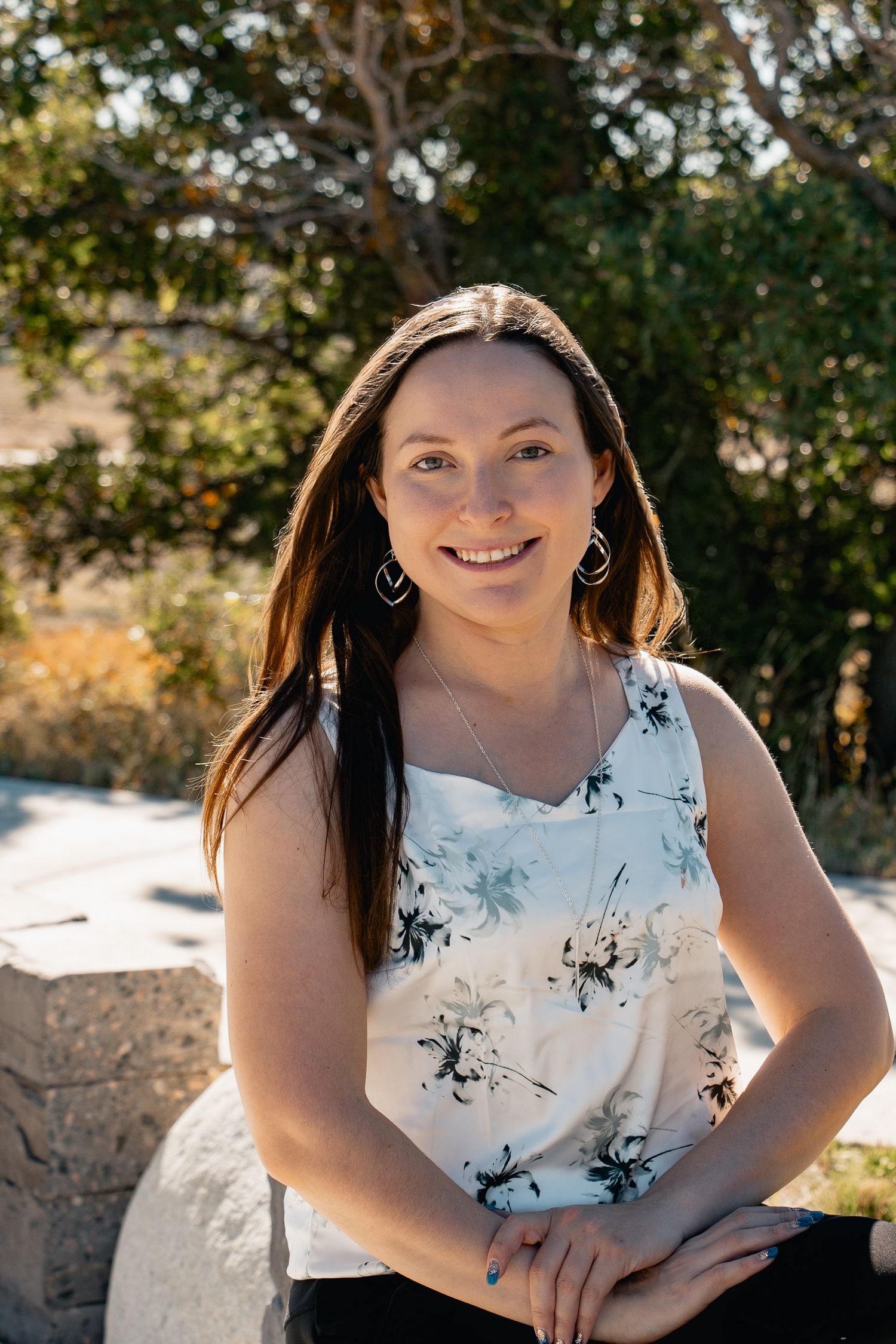 A person in a white floral dress poses outdoors against a natural background in warm sunlight.