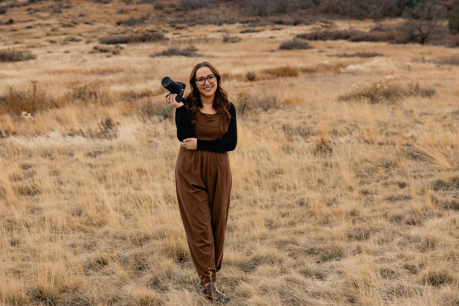 Woman in brown overalls holding a camera standing in a dry golden field with rocky hills in the background.