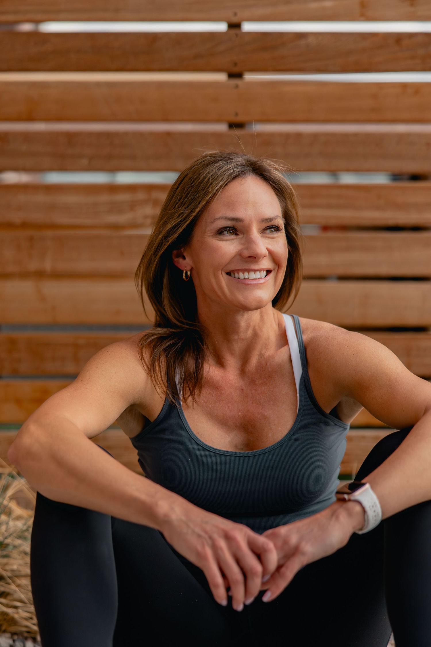 Smiling woman in athletic wear sitting relaxed against wooden slats, wearing a smartwatch, enjoying a fitness break outdoors.