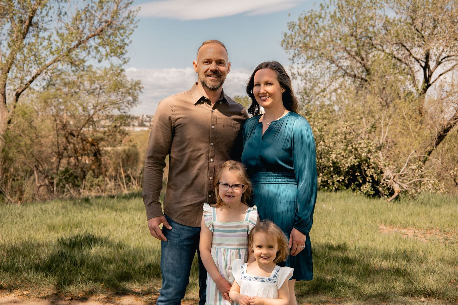 Happy family of four posing together outdoors on a sunny day with trees in the background.