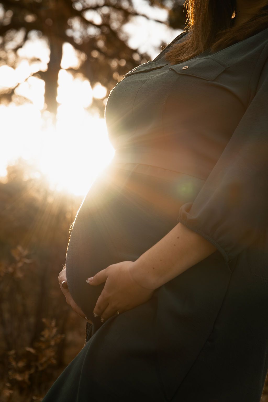 A pregnant woman's silhouette stands in golden sunlight while cradling her belly in a maternity photoshoot.