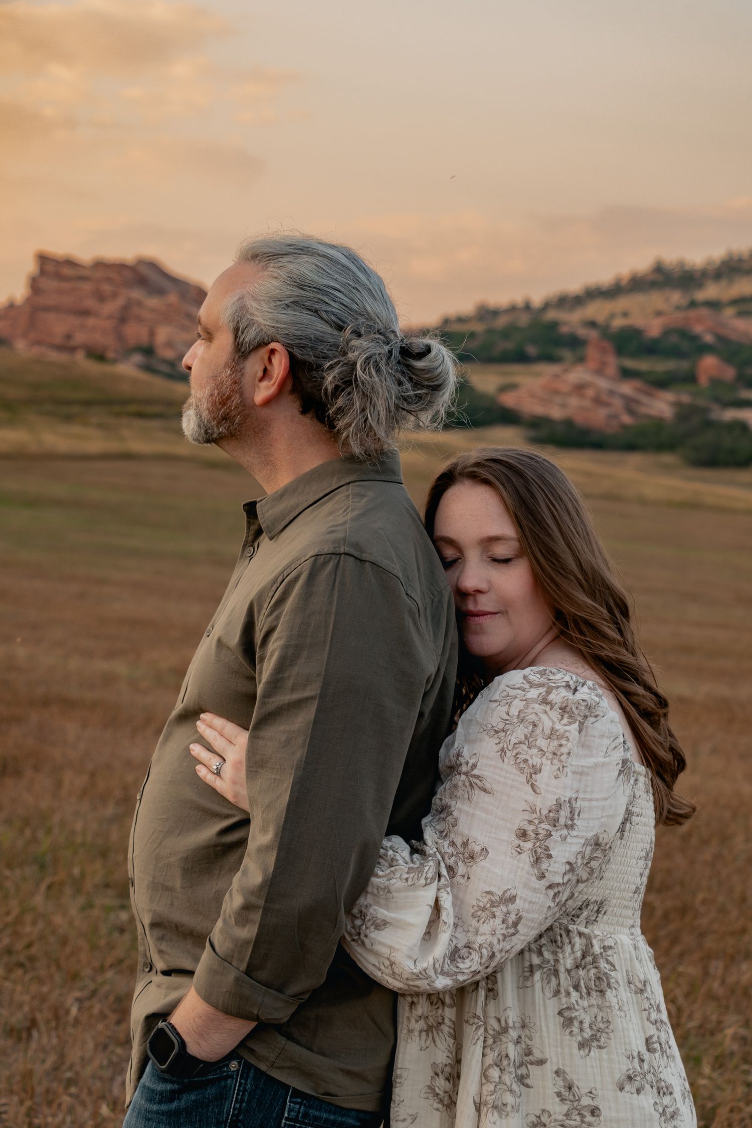Couple embraces in golden field at sunset, person wearing white lace dress and other in casual attire stands amid rolling hills.