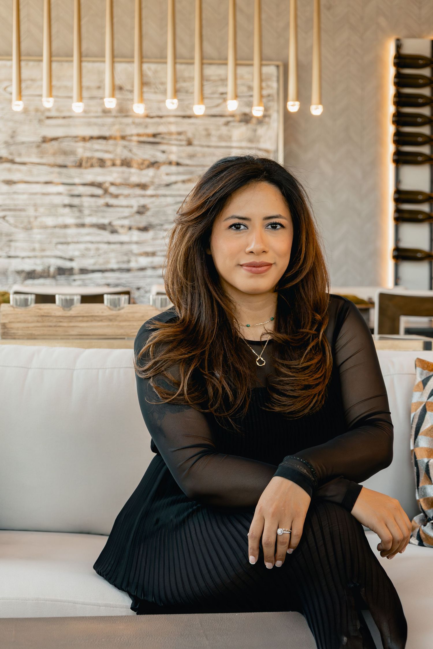 A person in black loungewear poses gracefully on a gray couch in an upscale interior setting with stone walls.