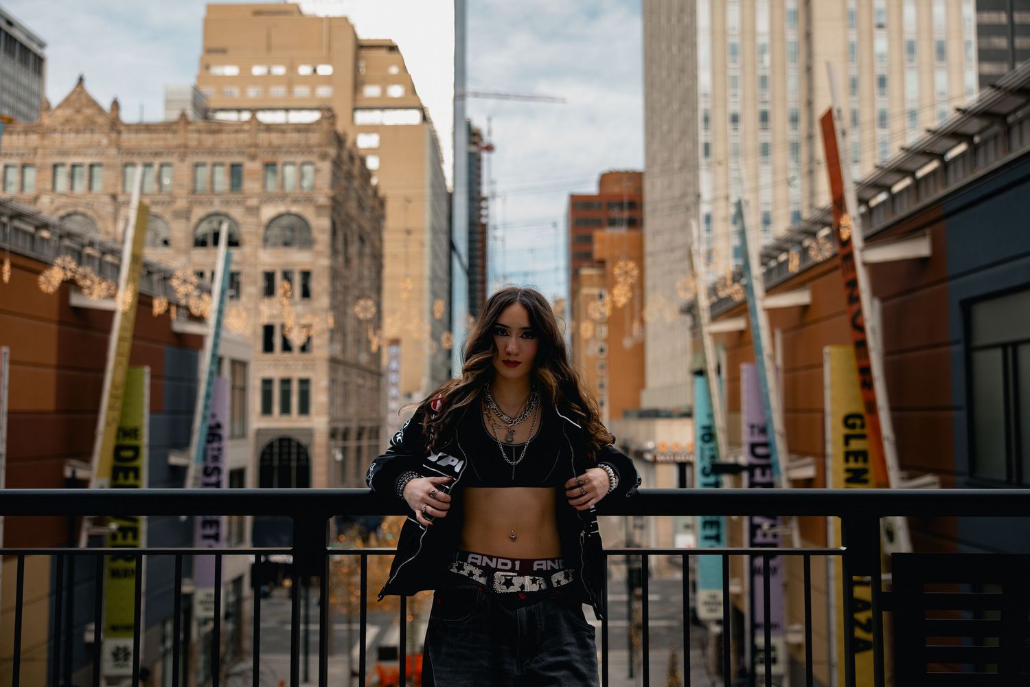 Young woman in crop top and black jacket poses on urban balcony with city buildings in background.