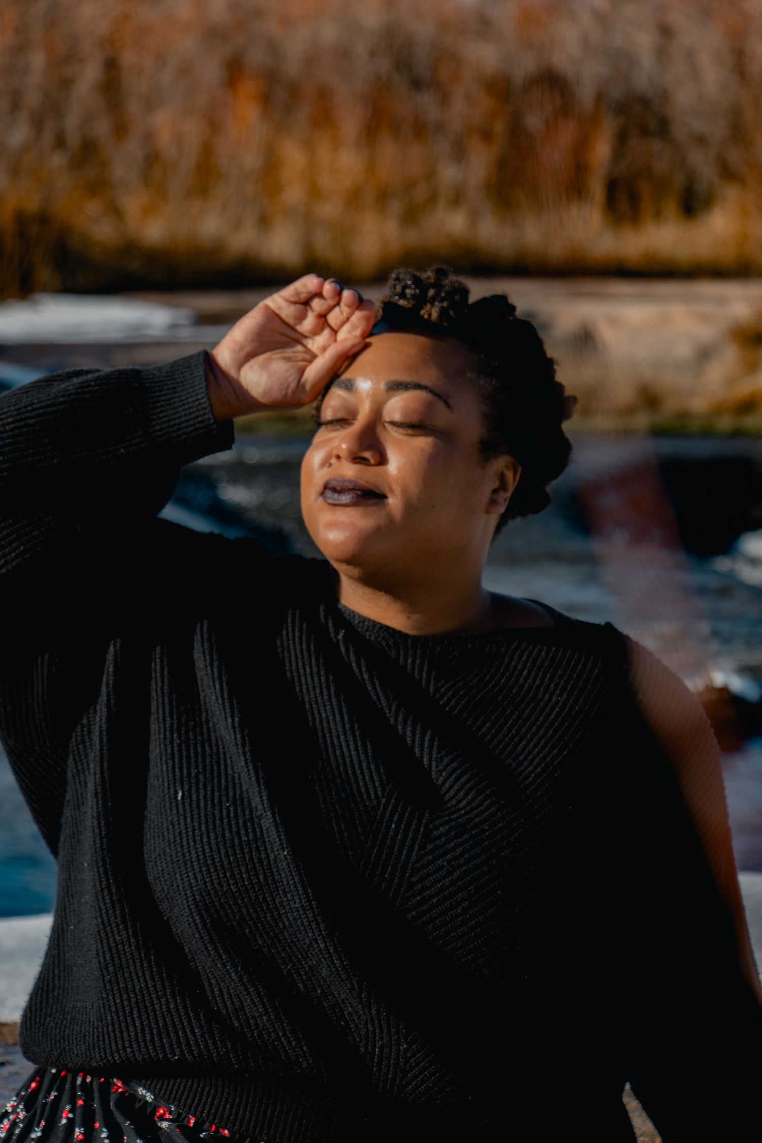 Woman in black sweater shielding eyes from bright sunlight outdoors near a pool on a sunny day.