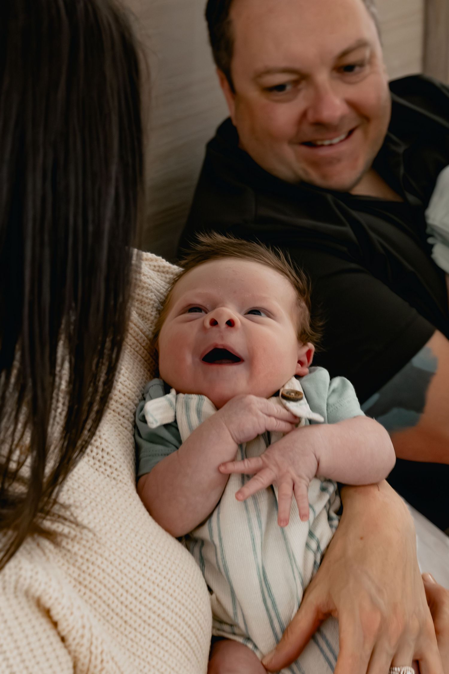 A happy newborn baby smiles while being held by parents, with dad smiling warmly in the background.