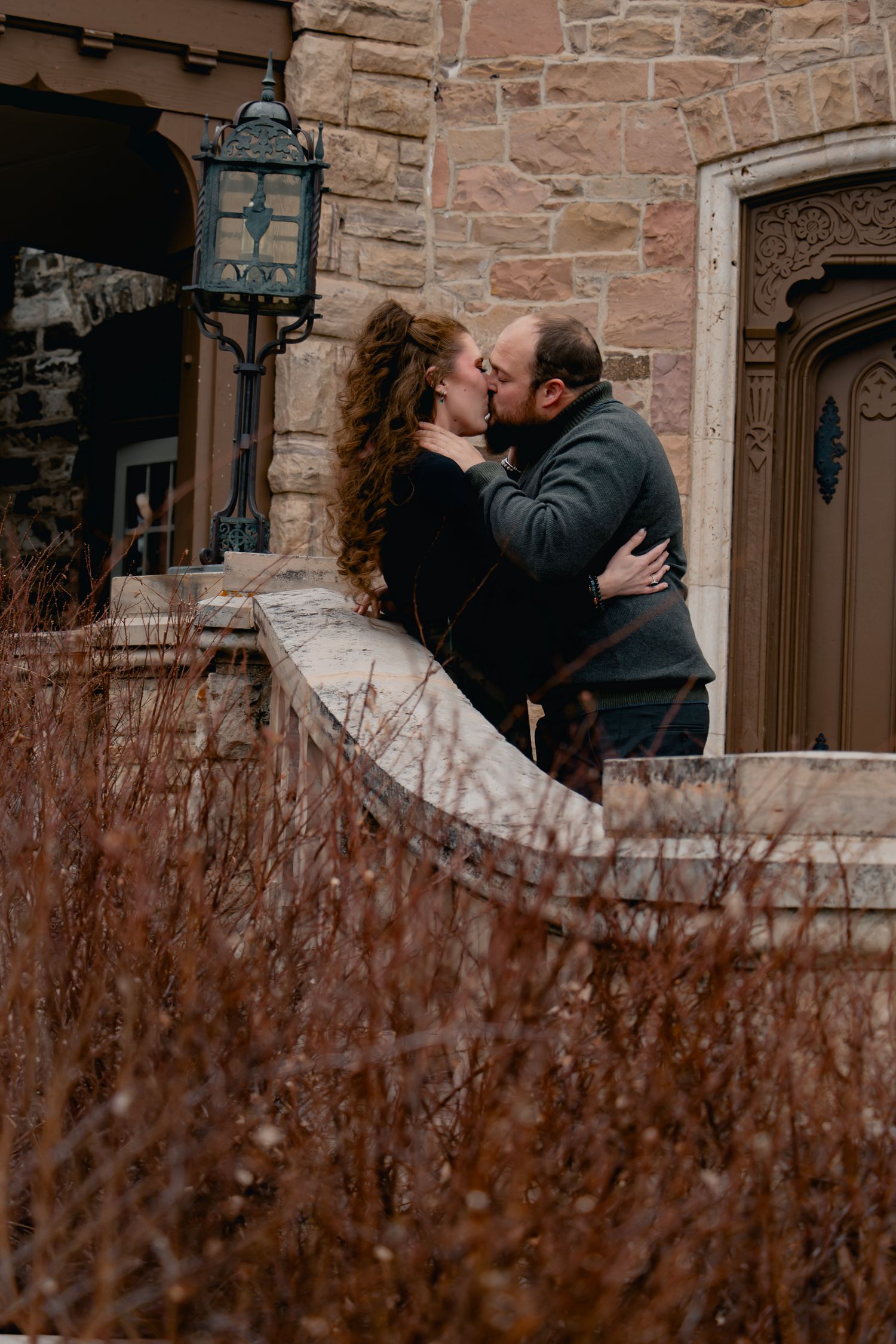 A romantic couple shares a tender kiss against a rustic stone wall backdrop with winter foliage in the foreground.