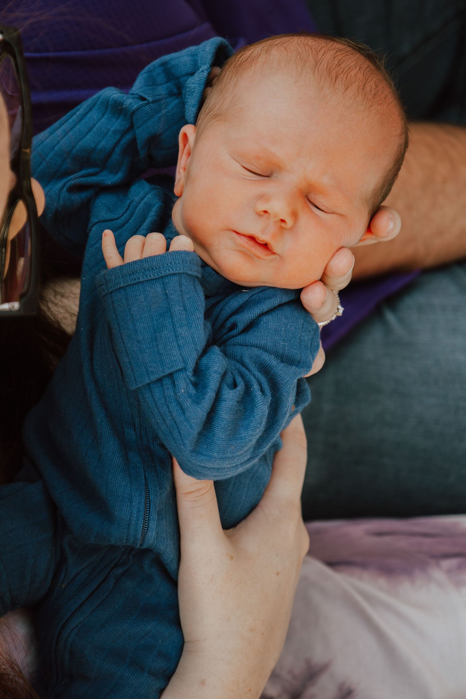 A sleeping baby in a blue outfit rests peacefully while being cradled in someone's arms.