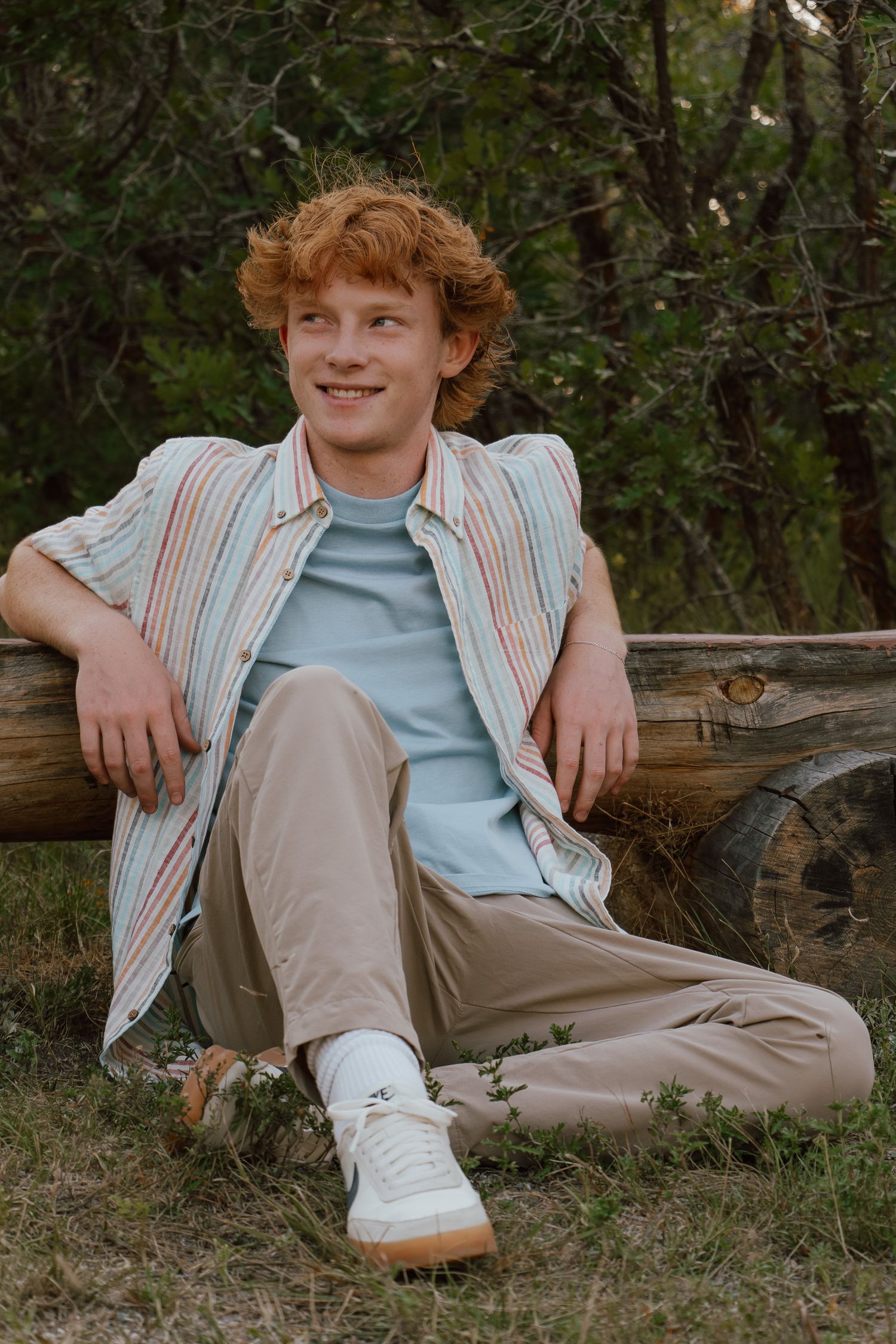 A casual portrait of someone lounging outdoors in a striped button-down shirt, khaki pants and white sneakers.