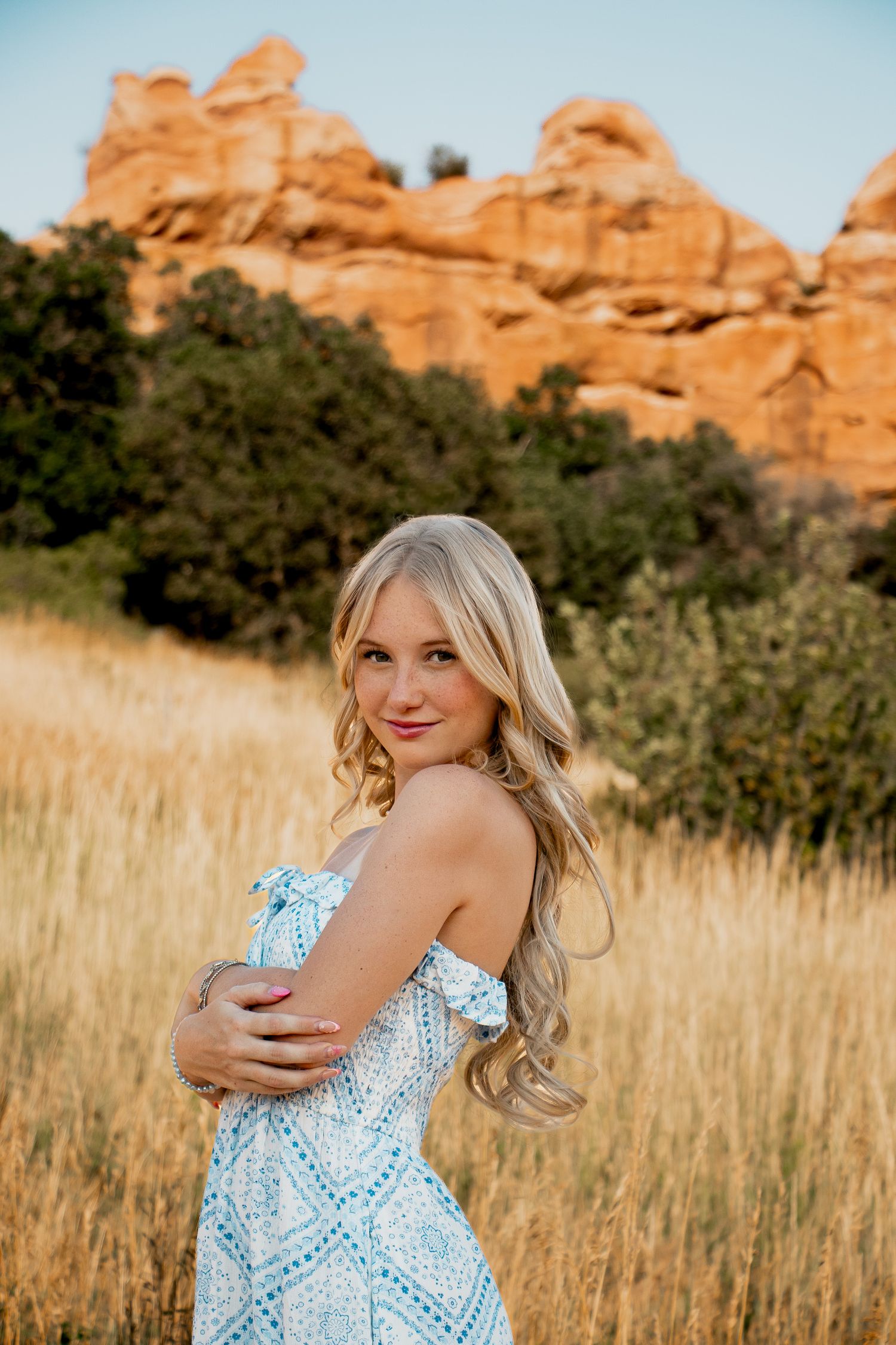 Person poses in front of red rock formations while wearing a blue and white patterned dress in a grassy field.