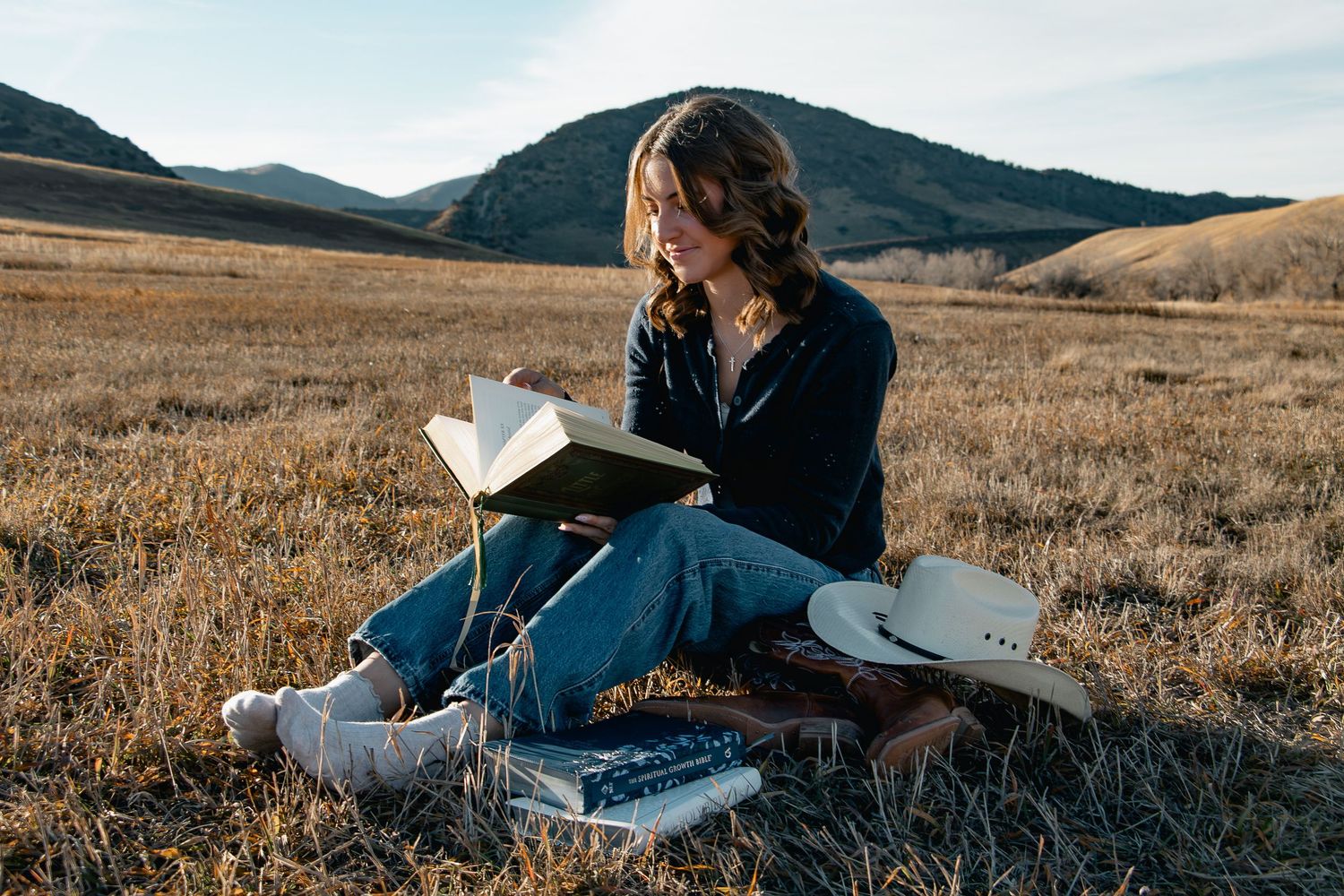 Person in denim sits on grassy hillside reading a book against backdrop of mountains during golden hour.