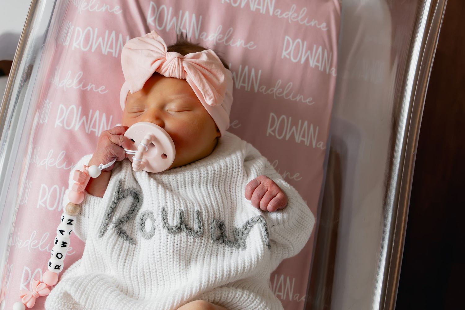 A newborn baby wearing a white knit sweater and pink bow headband rests on a pink personalized name blanket.
