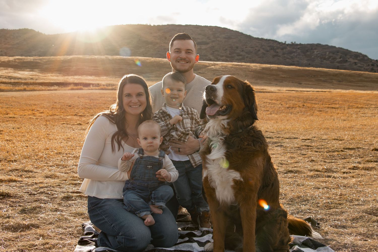 A family and their dog sit together in a golden field during sunset as warm sunlight streams through the mountains.