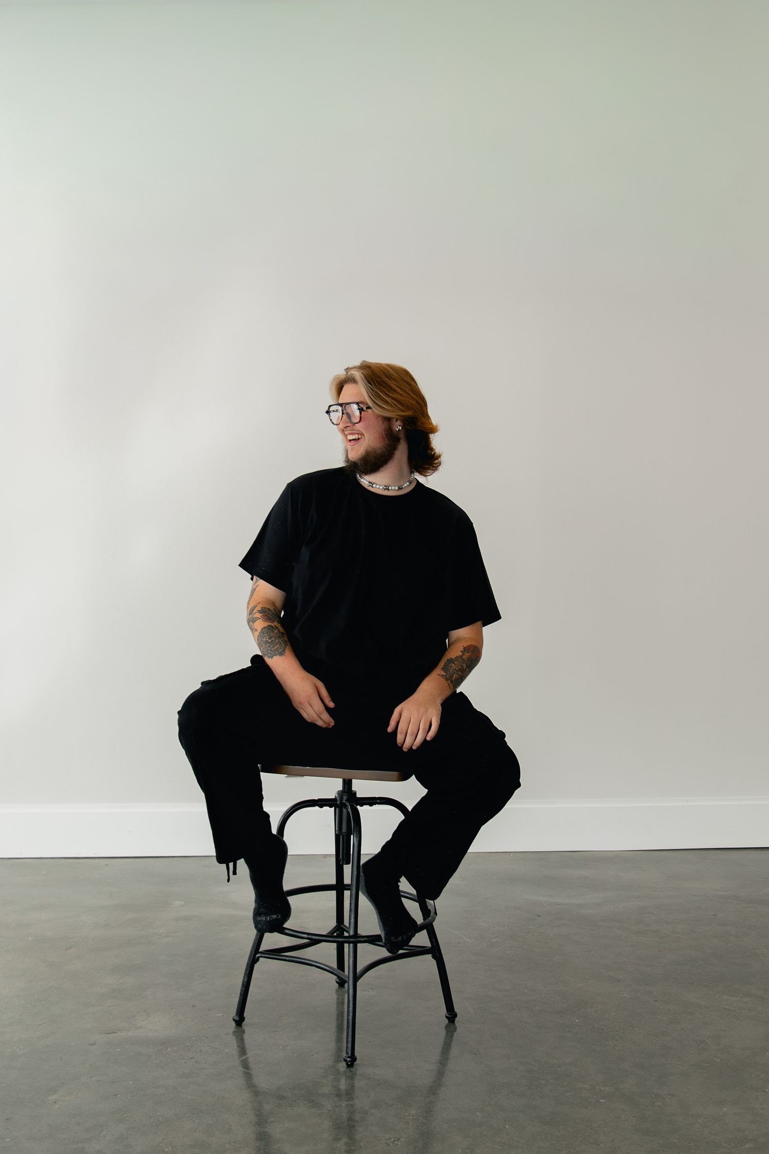A person in black clothing sits casually on a dark chair against a white wall in a minimalist setting.