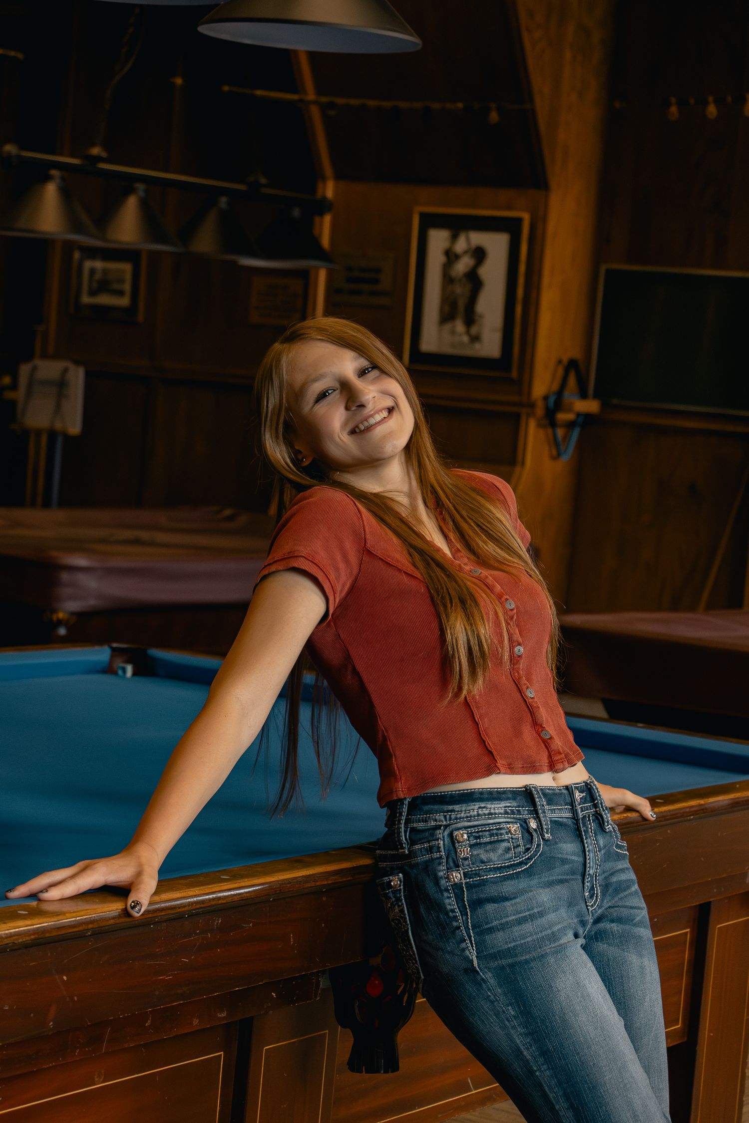 A person in a red shirt and jeans leans casually against a pool table in a dimly lit billiards room.