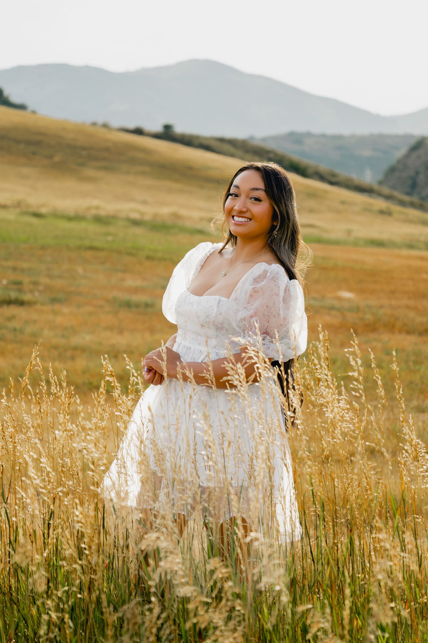 A flowing white dress twirls through a golden field of tall grass with rolling hills in the background during sunset.