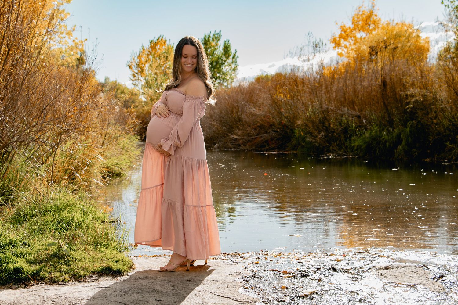 A figure in a flowing peach dress stands gracefully by a serene autumn riverside with golden foliage.