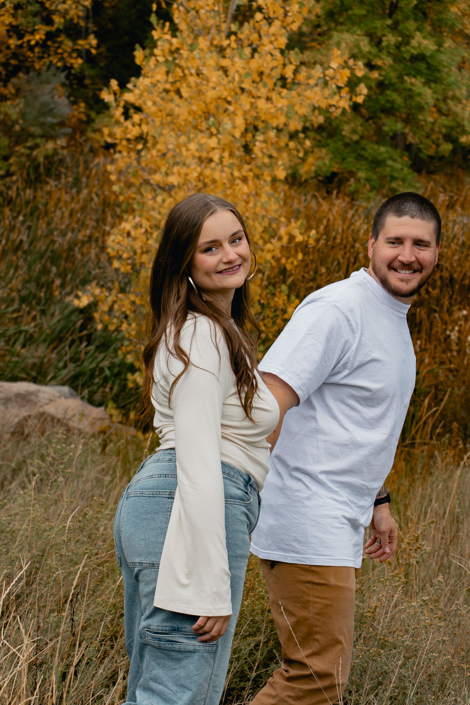 A couple shares playful moments in autumn scenery with golden leaves and tall grass in the background.