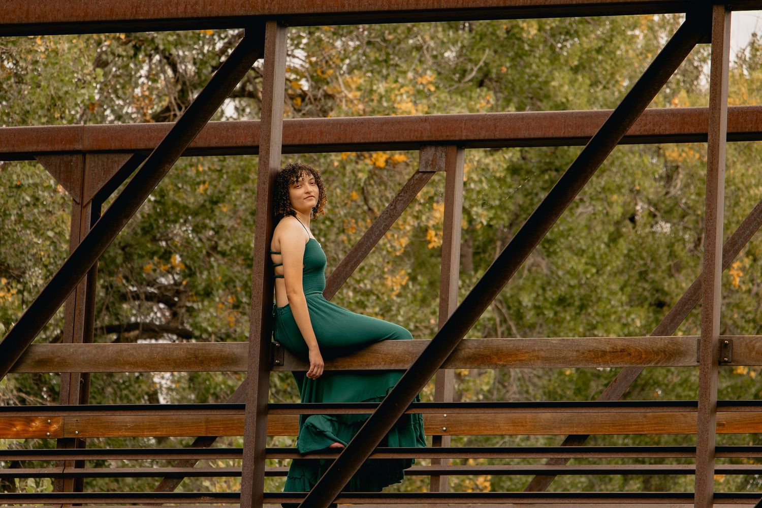 A woman in an emerald green dress poses gracefully on a rustic wooden bridge in a natural outdoor setting.