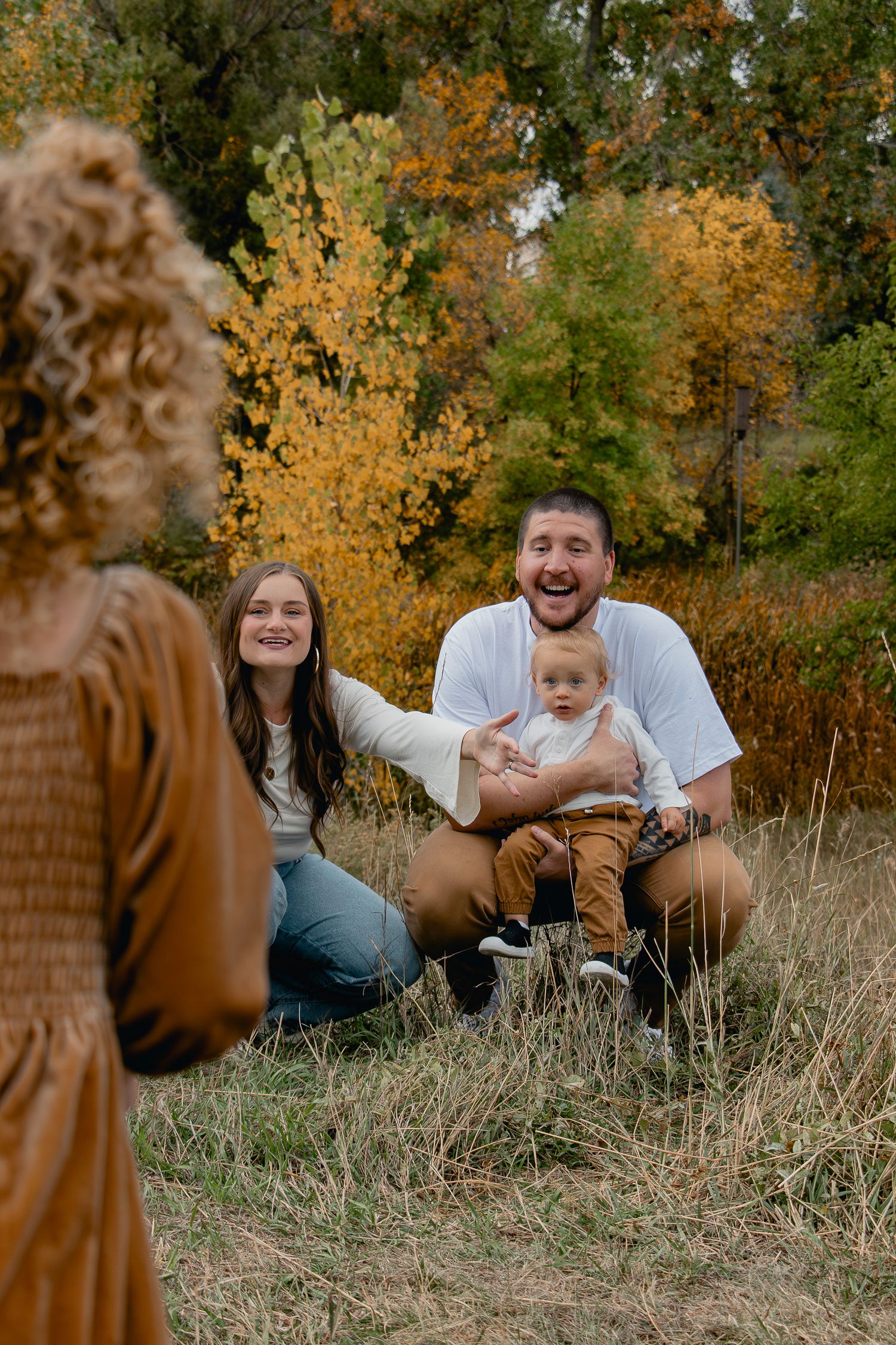 A family enjoys a candid moment together while sitting in autumn grass with golden foliage in the background.