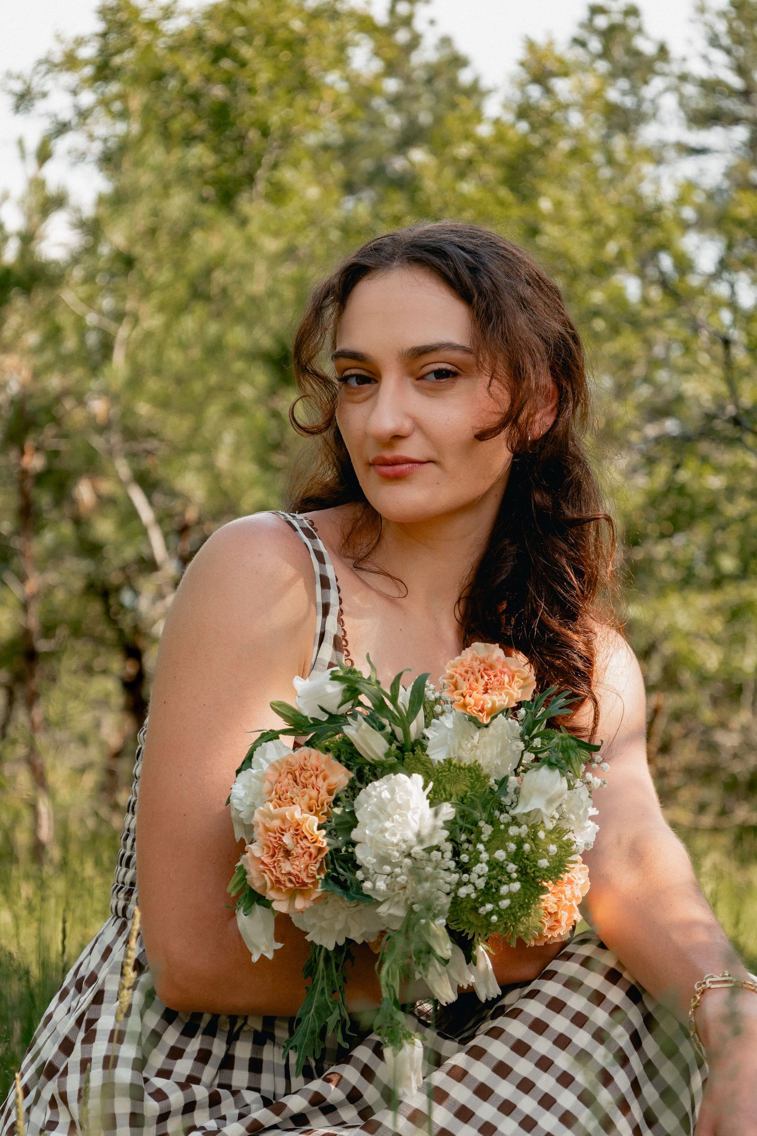 Someone in a gingham dress holds a beautiful bouquet of peach and white flowers against a natural outdoor background.