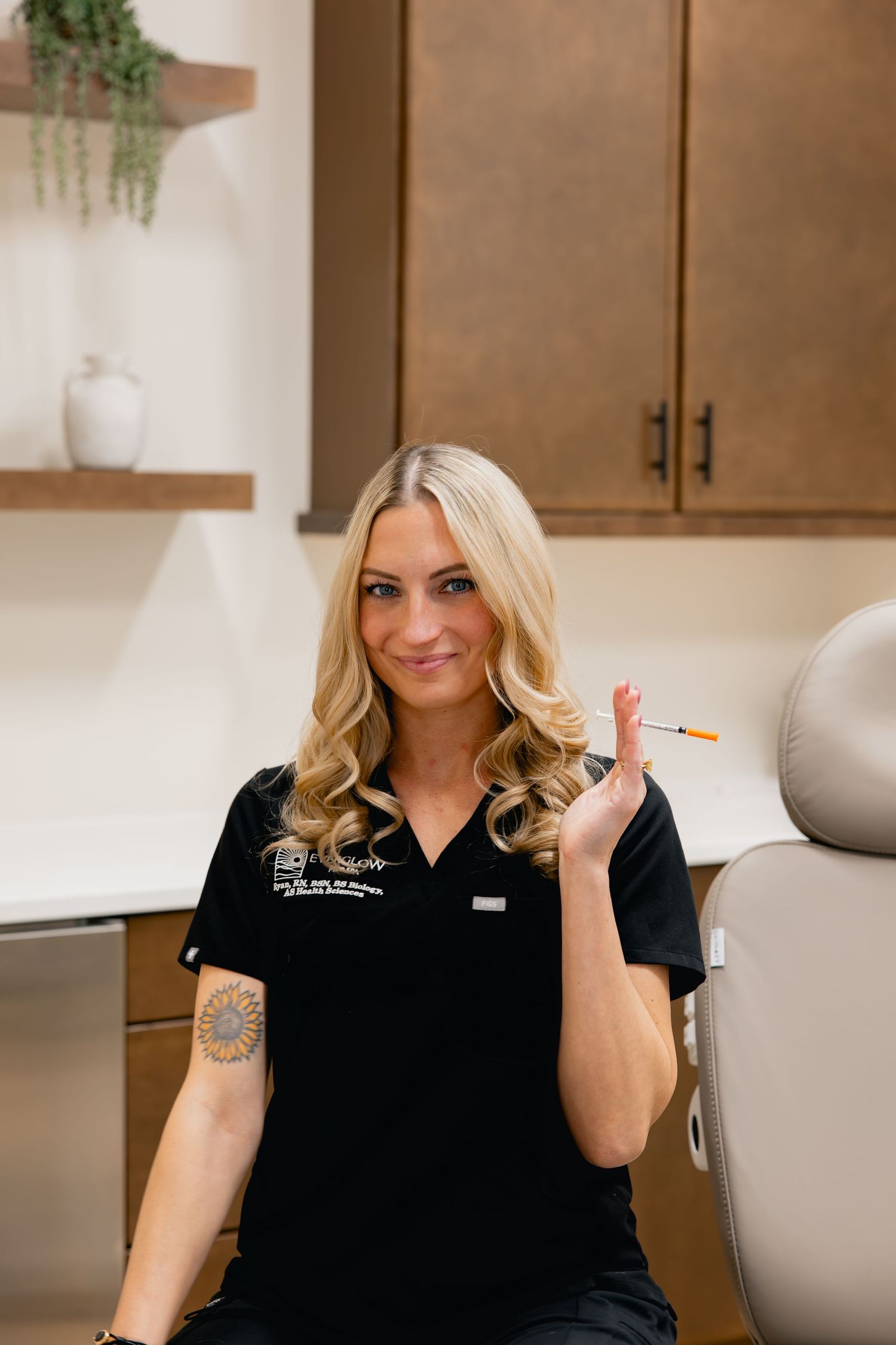 A blonde female medical professional in a black uniform holds a syringe in a modern clinical treatment room.