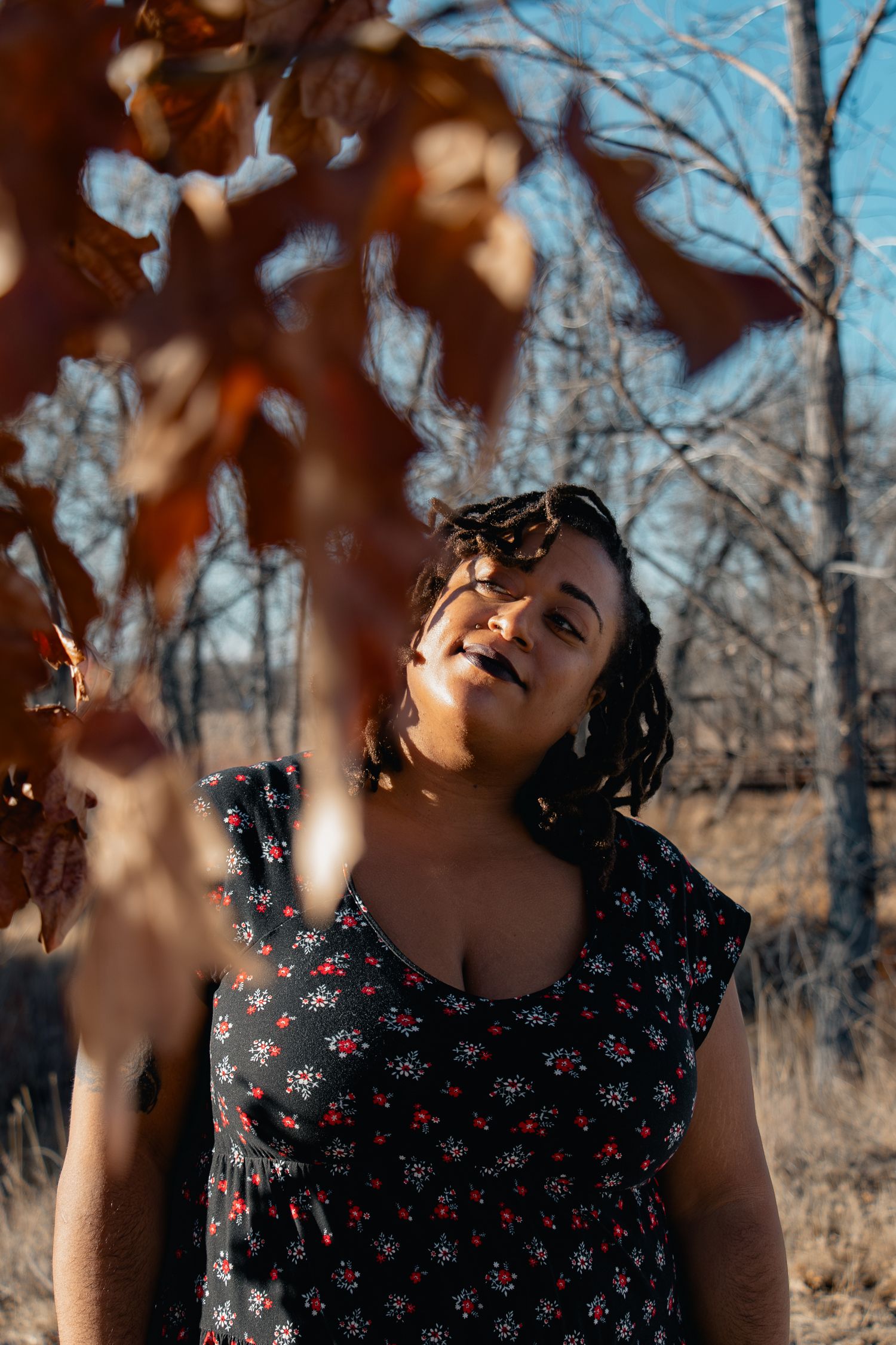 Woman in floral dress gazing upward among autumn leaves and bare trees in golden sunlight.