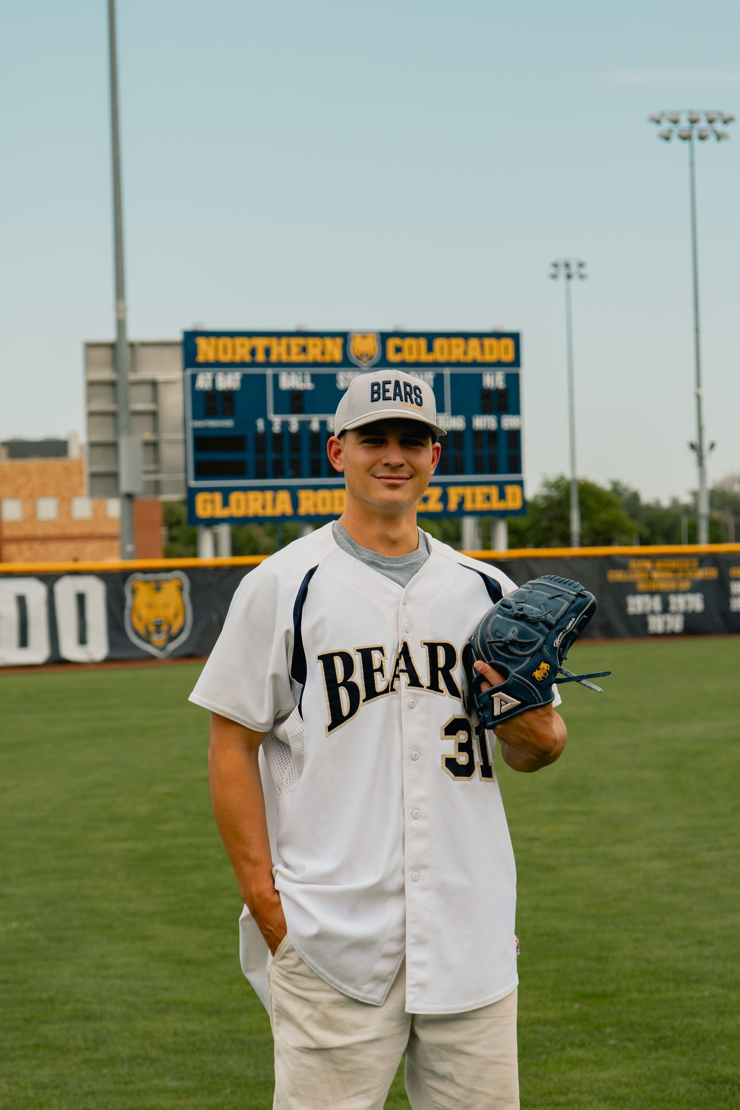 Baseball player in white Bears jersey stands on field with glove at Northern Colorado baseball stadium scoreboard.