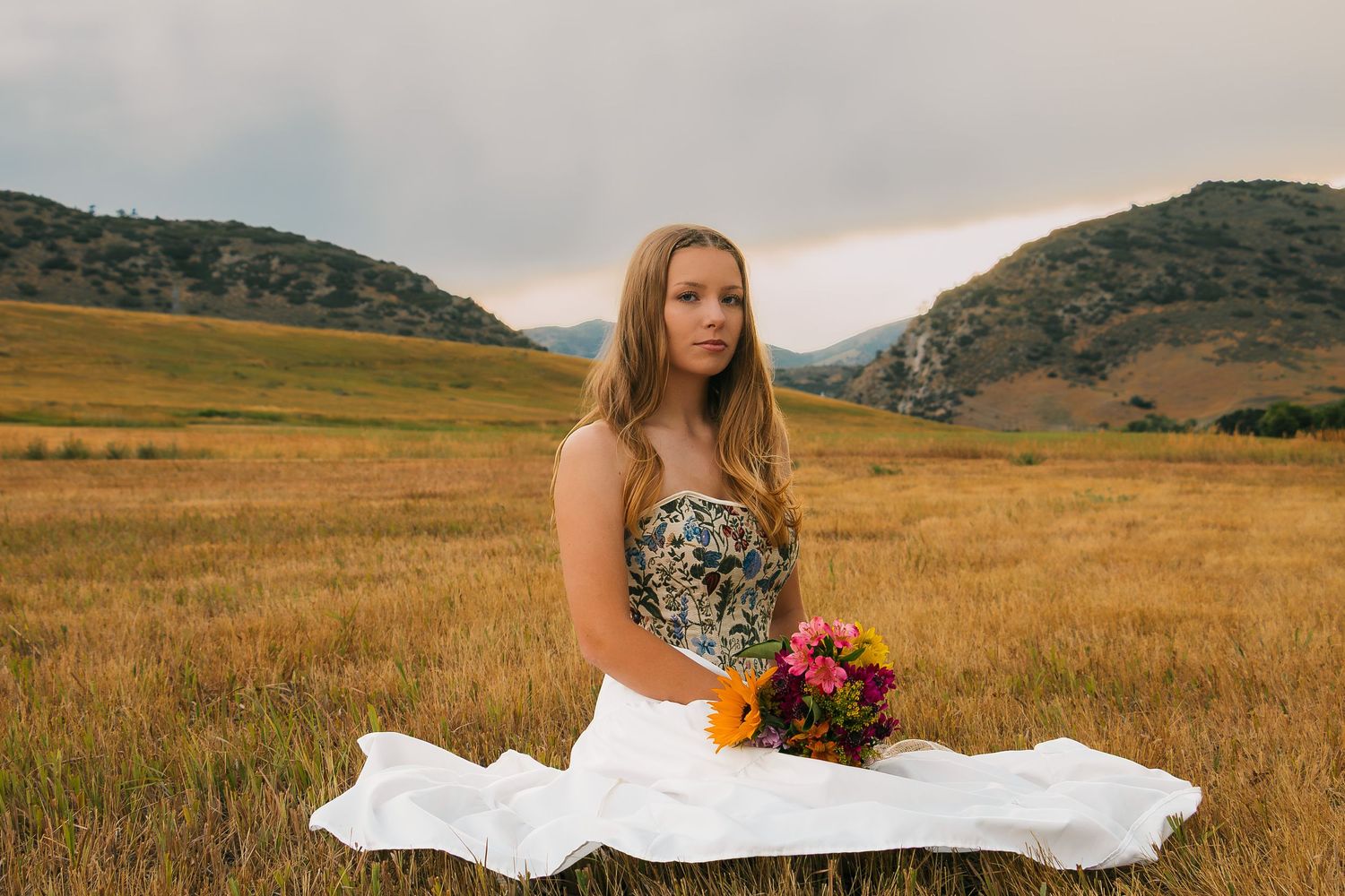 A white dress flows gracefully in the wind as someone stands in a golden meadow with mountain views at sunset.