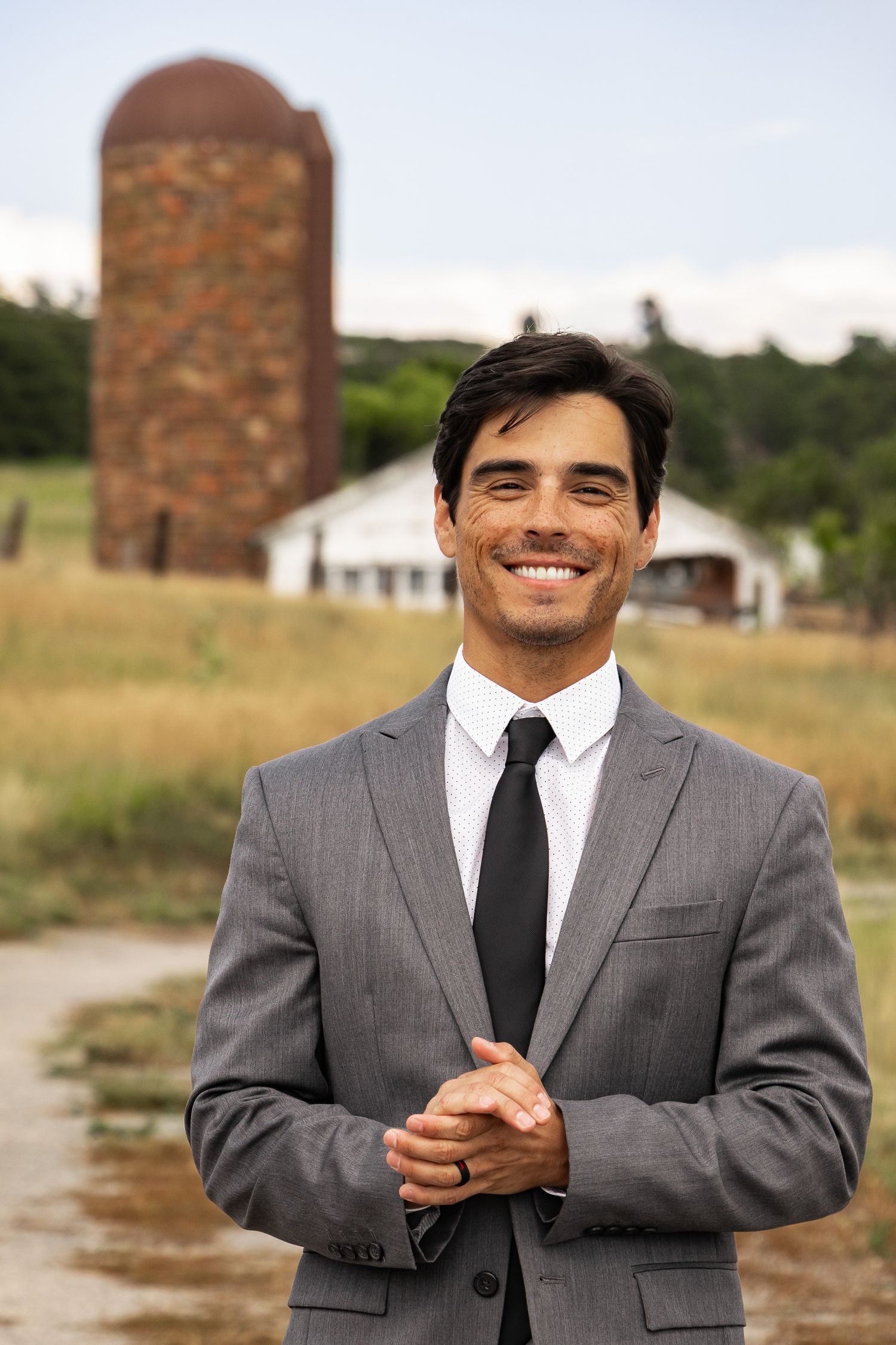 Professional in a grey suit and black tie smiles while standing in front of a rustic barn and silo in a rural setting.