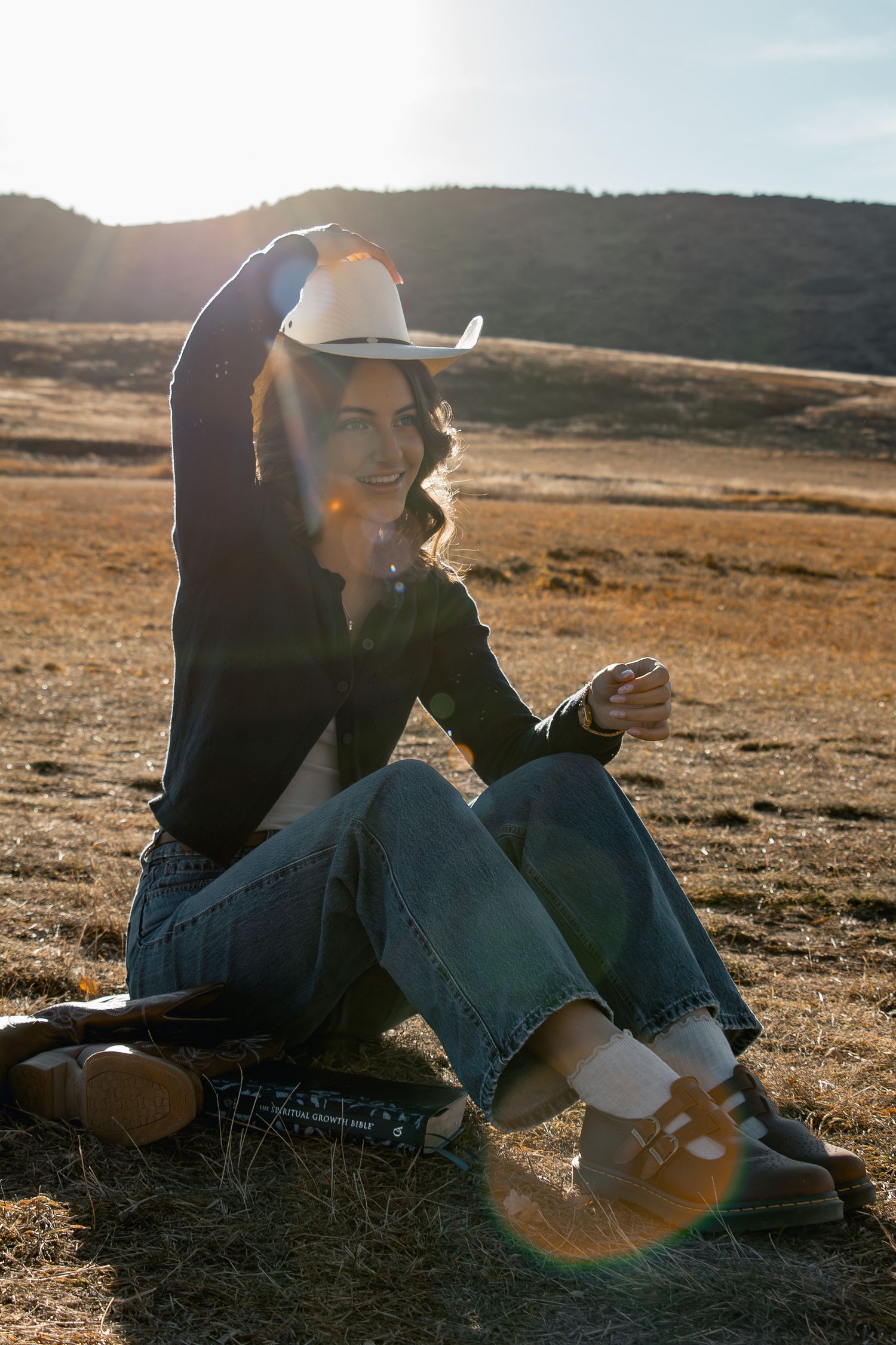 Person in cowboy hat and jeans sitting on ground during golden hour with mountains in background.