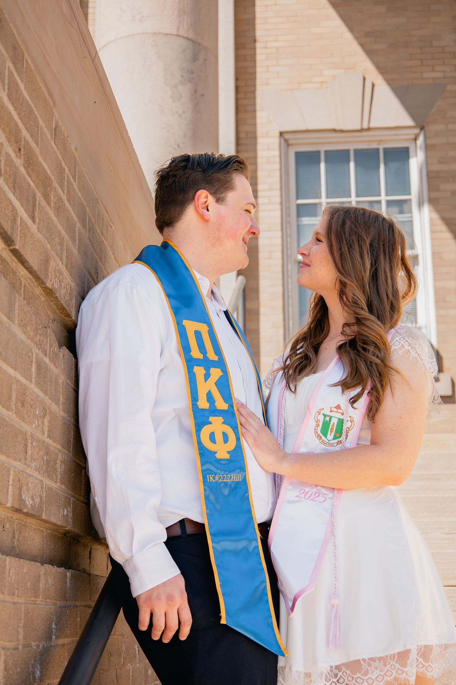 A couple in white outfits and Greek letter organization stoles gaze at each other against a brick wall background.