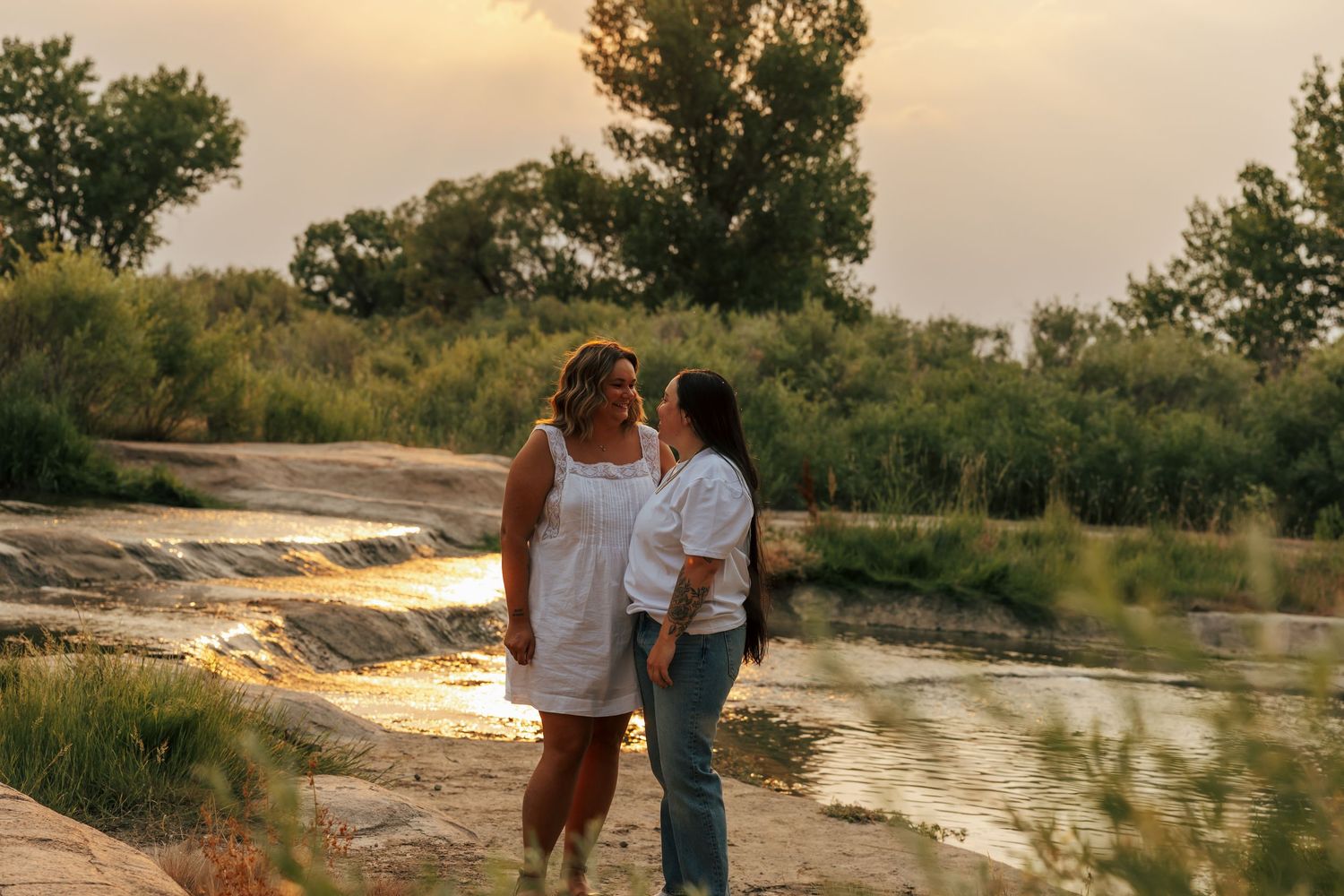 Two women share a tender moment by a serene river at golden hour, surrounded by lush greenery and warm sunset light.