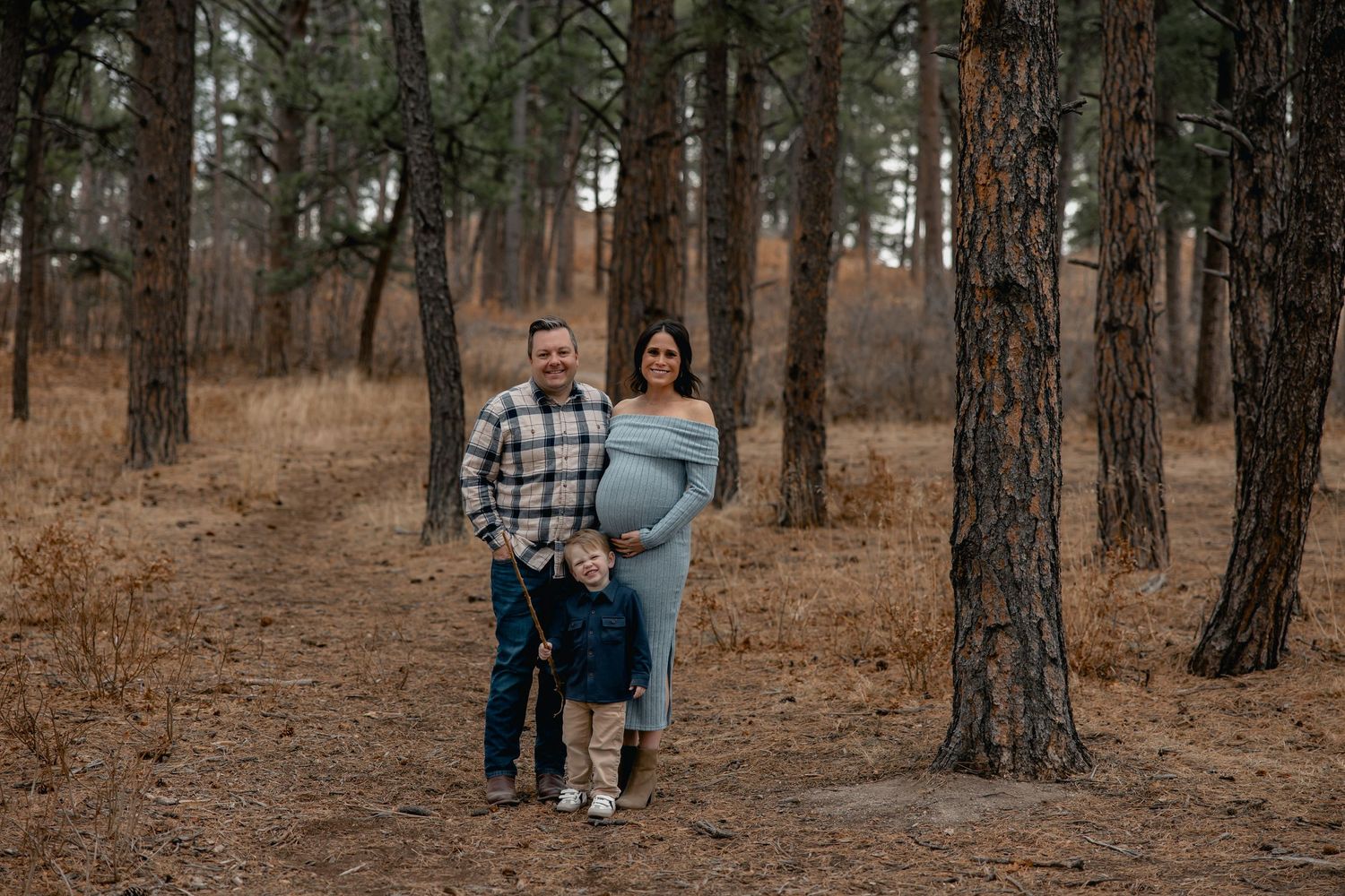 Pregnant couple with toddler posing on forest path among tall pine trees during maternity photo session.