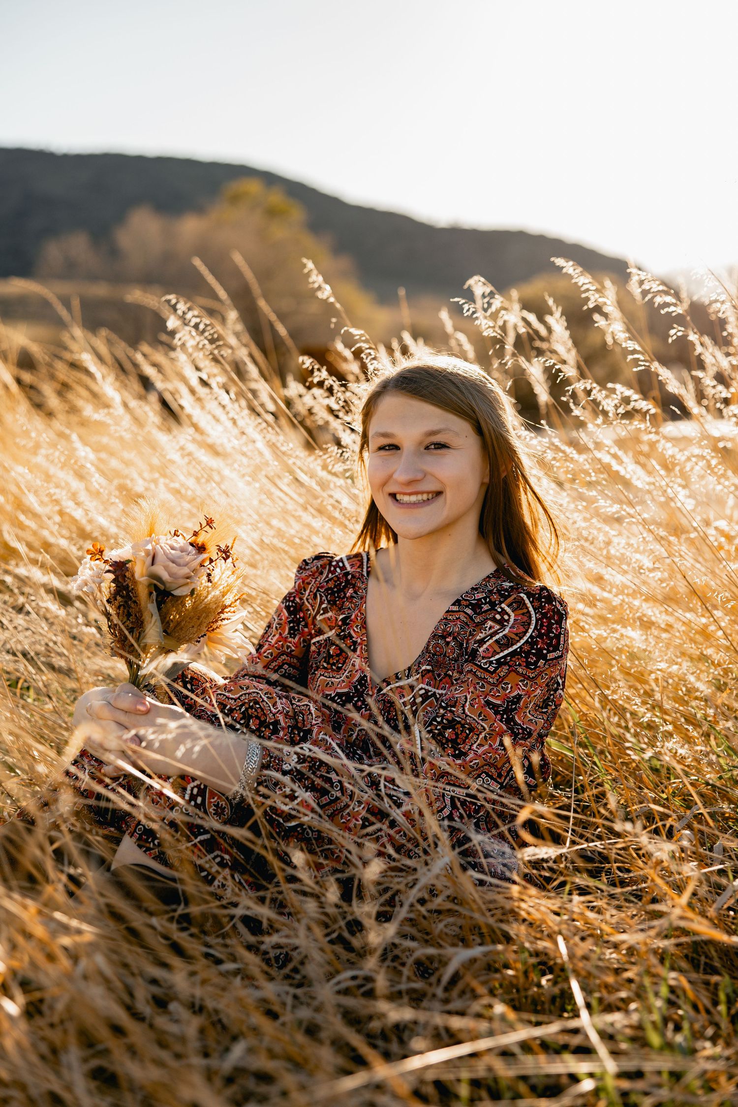 Smiling woman in paisley dress sitting in golden field grass during sunset with mountain backdrop.