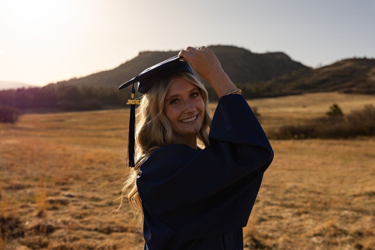 Young woman in graduation cap and gown smiling outdoors in a golden field with mountains at sunset.
