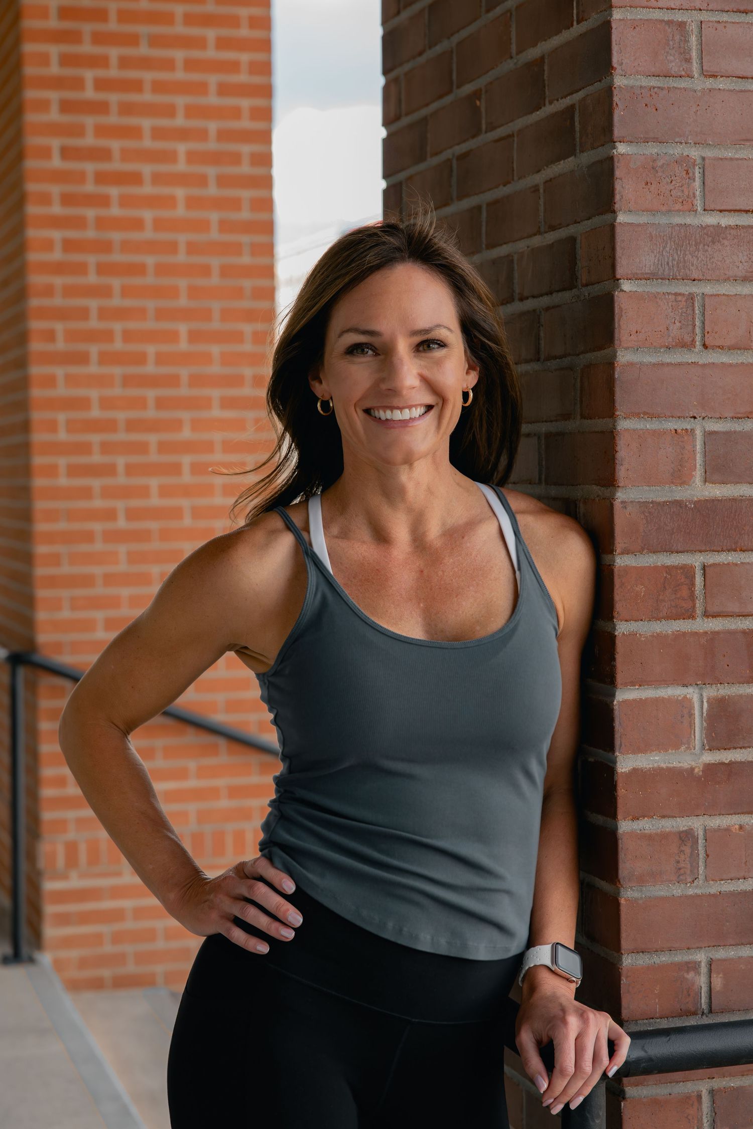Smiling woman in gray racerback tank and black leggings leaning against brick wall outdoors.
