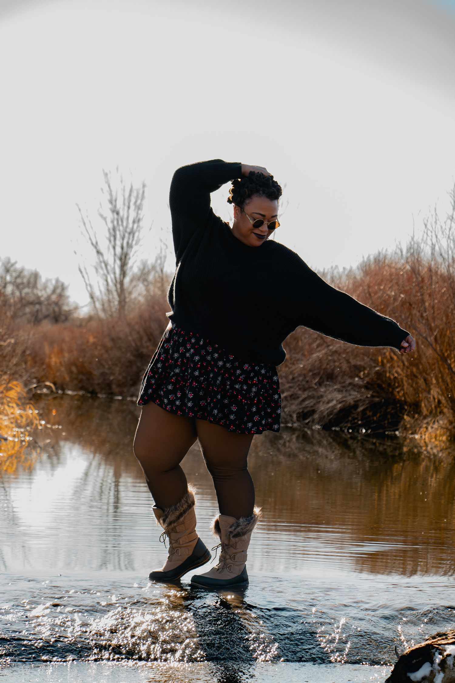 Woman in black sweater, floral skirt, and boots splashing in a shallow river during autumn.