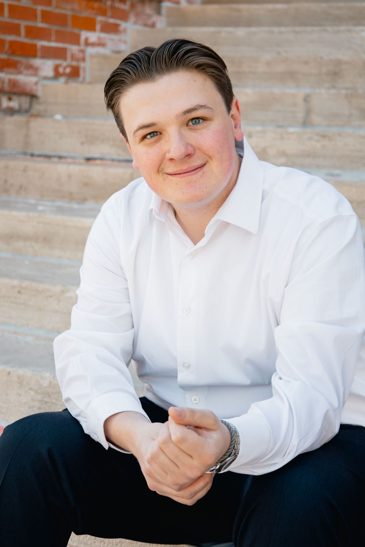 Professional portrait of a person in a white dress shirt sitting casually on concrete steps with brick wall background.