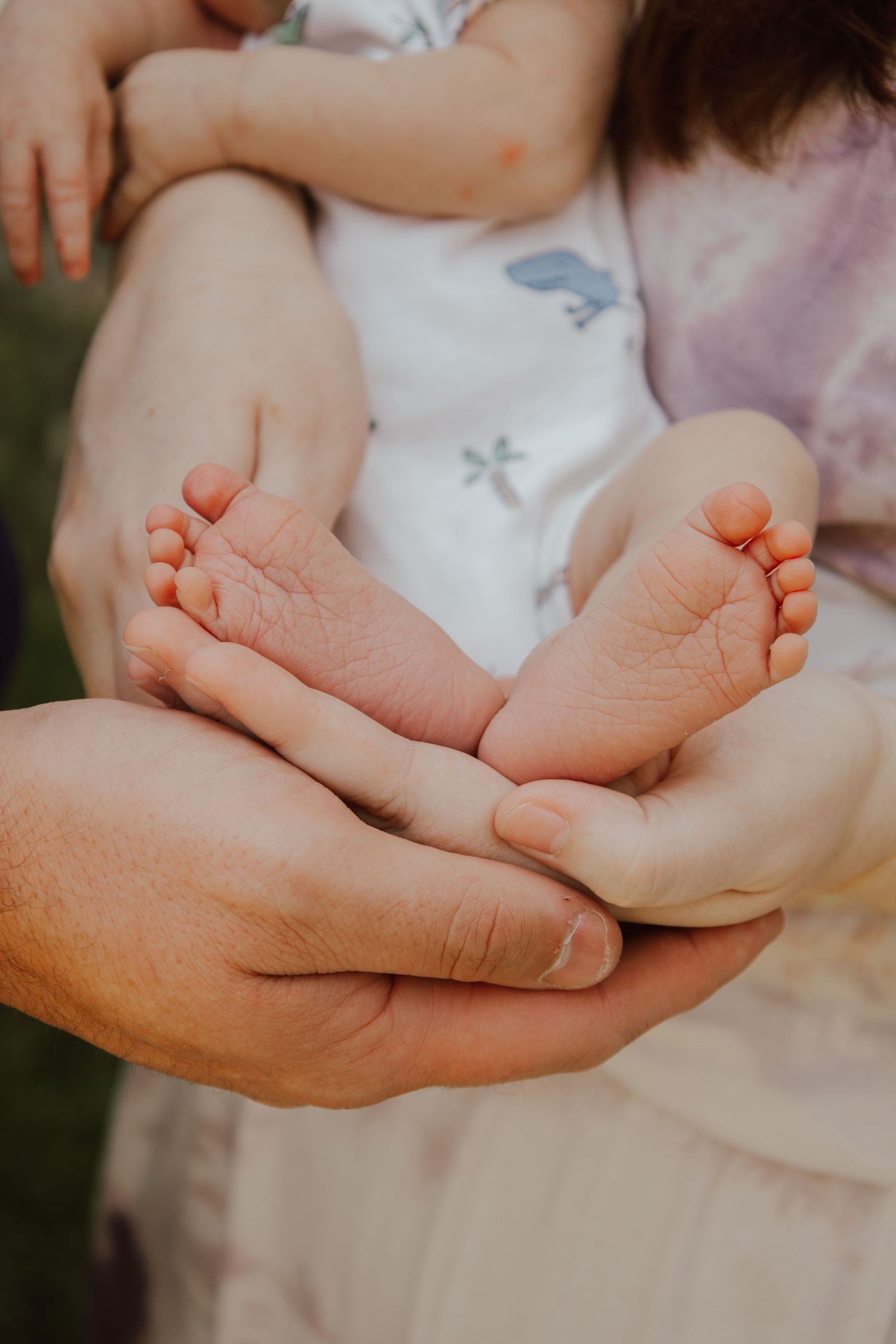 Gentle hands cradle tiny newborn baby feet in white clothing against soft blurred background.
