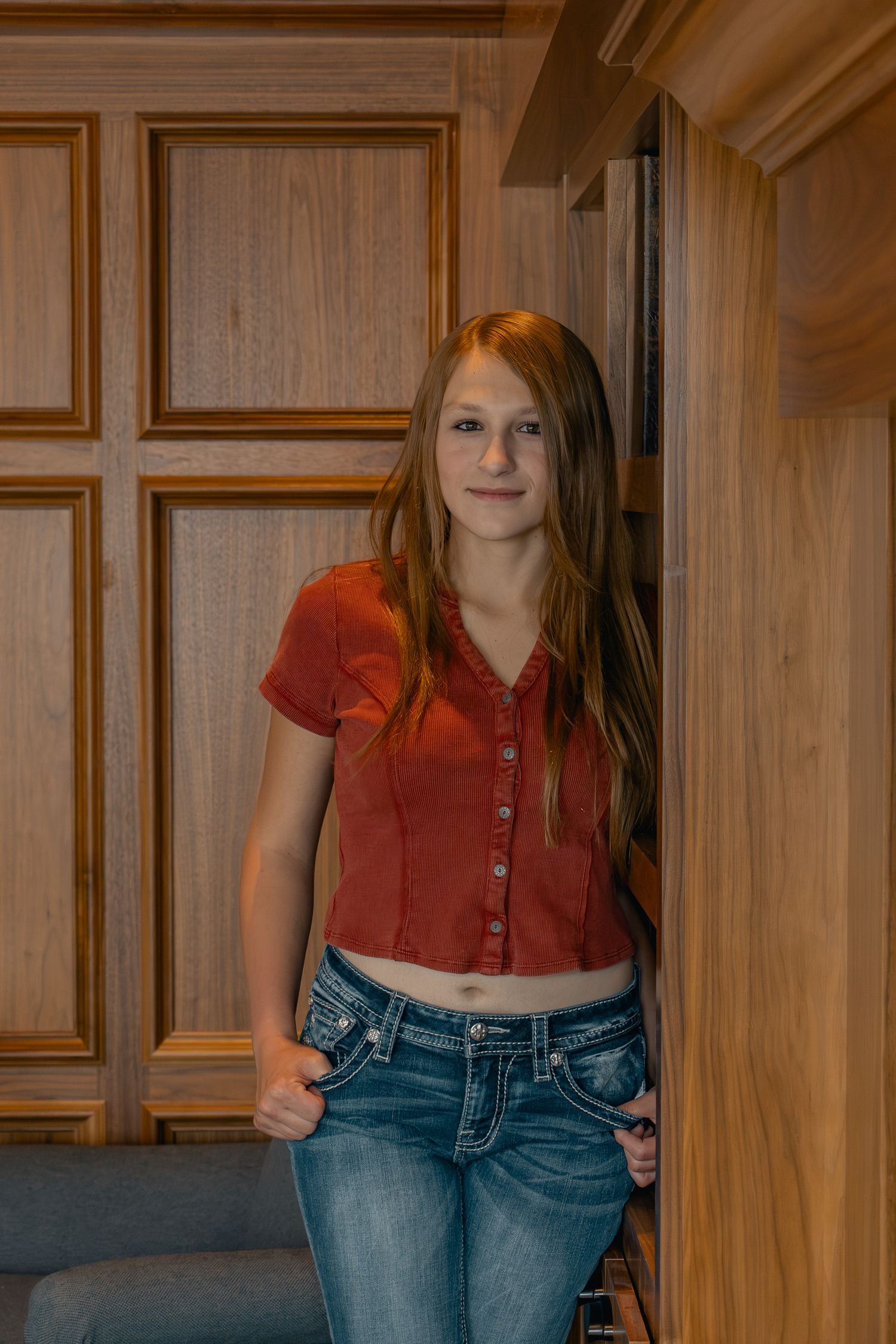 A person wearing a red crop top and blue jeans poses in front of wooden paneled walls.