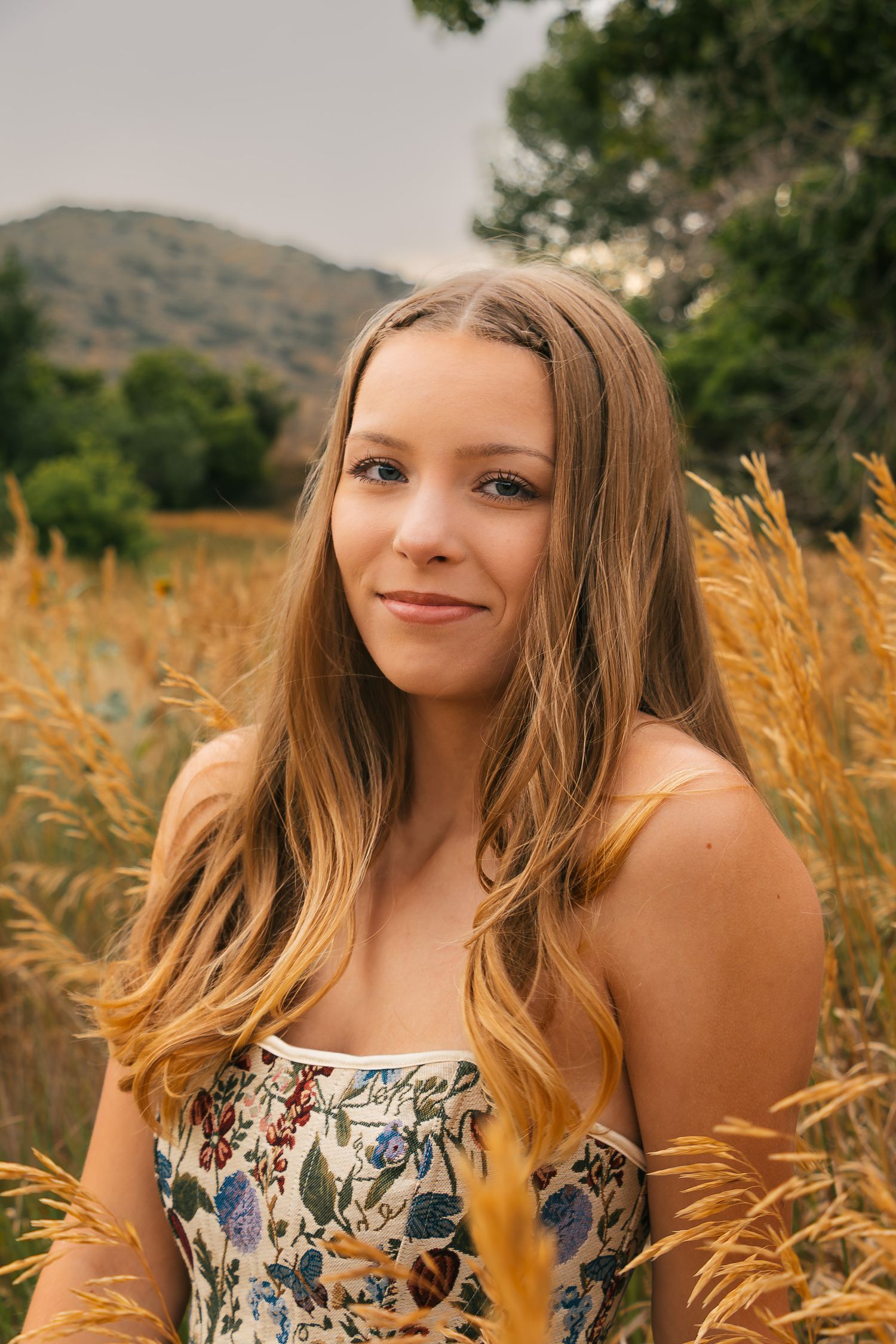 Portrait photo in a golden wheat field with mountains and trees in the background on a sunny day.