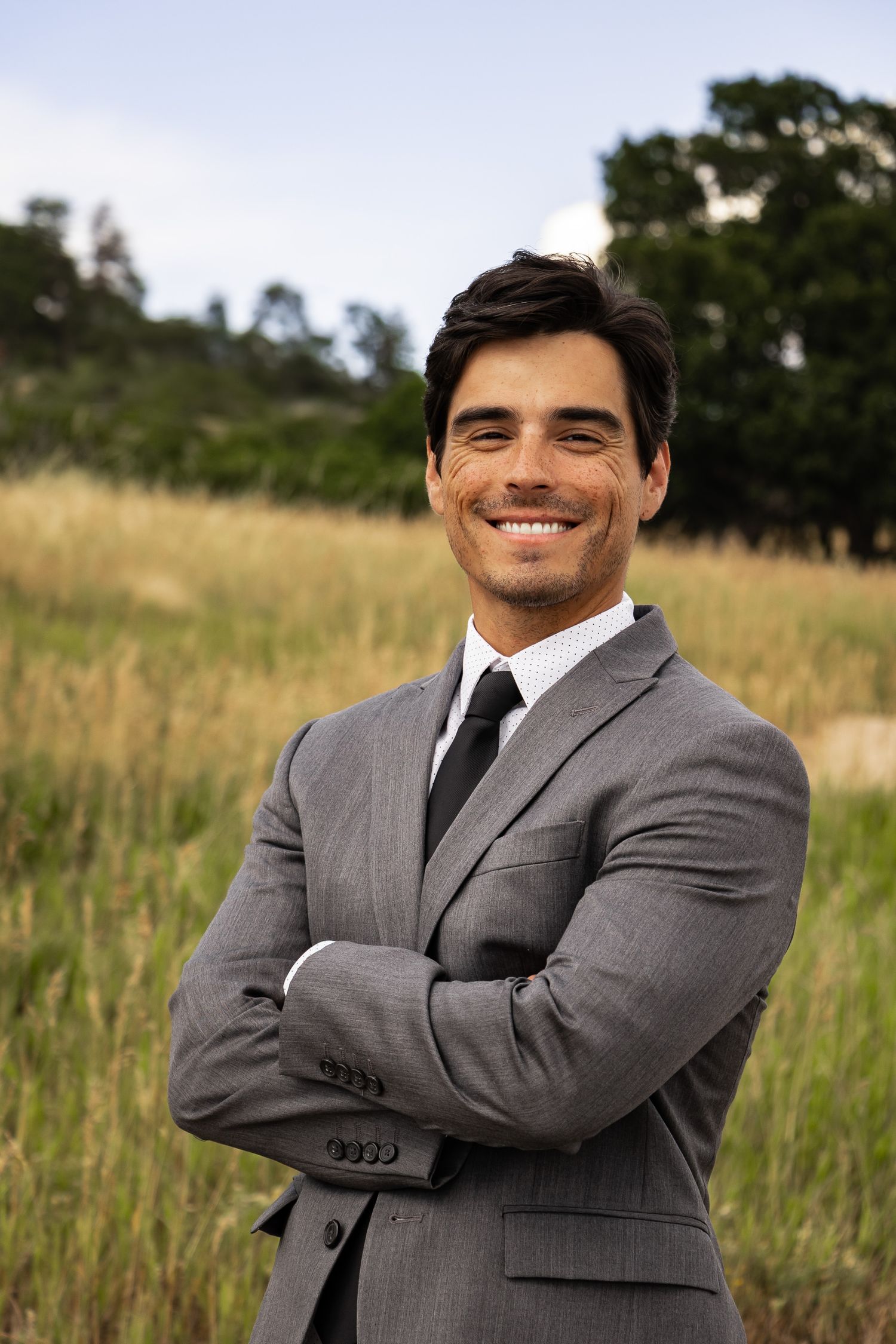 Professional in a gray suit poses confidently against a natural outdoor field background.