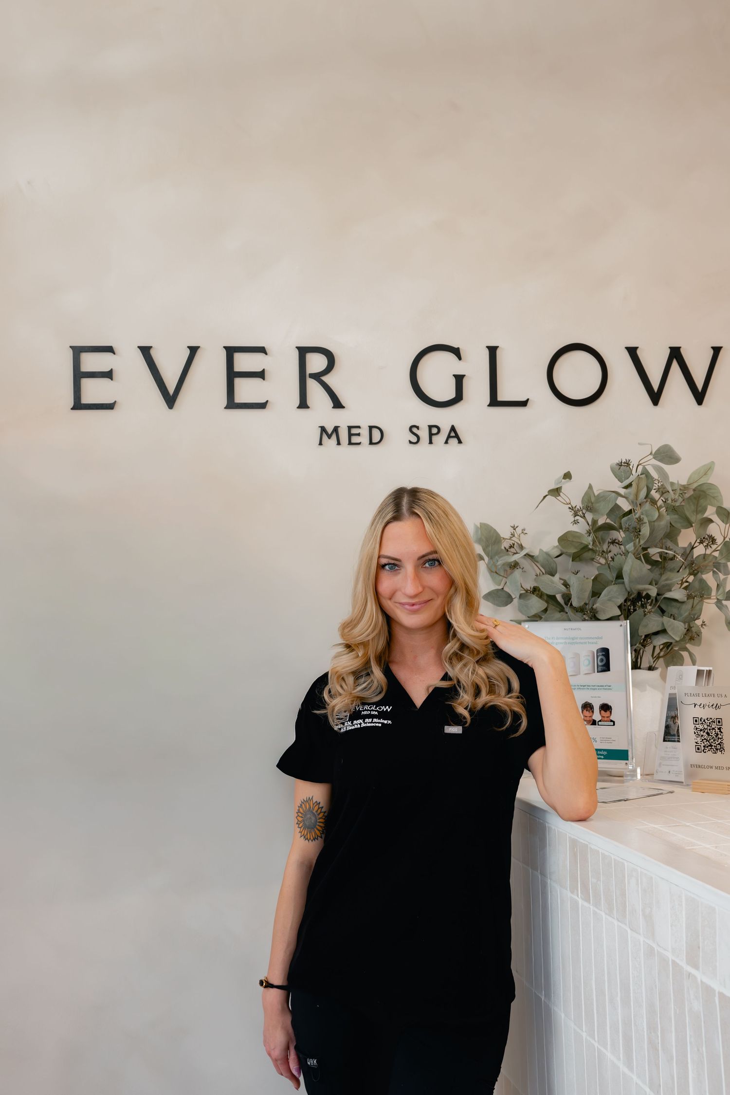 Smiling blonde woman in black scrubs poses at Ever Glow Med Spa reception desk with greenery decor.