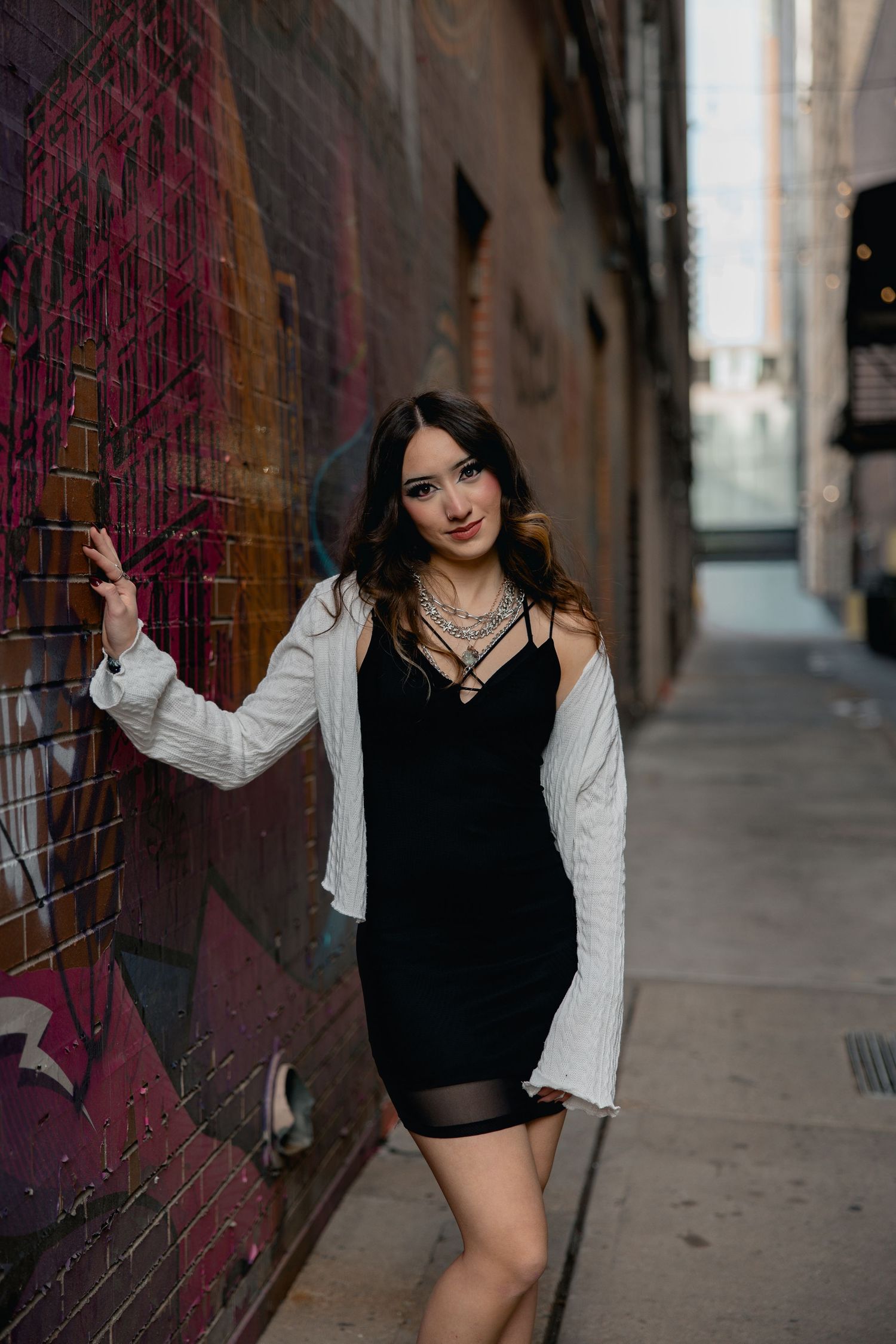 Young woman in black mini dress and white cardigan posing in urban alley with colorful graffiti wall.